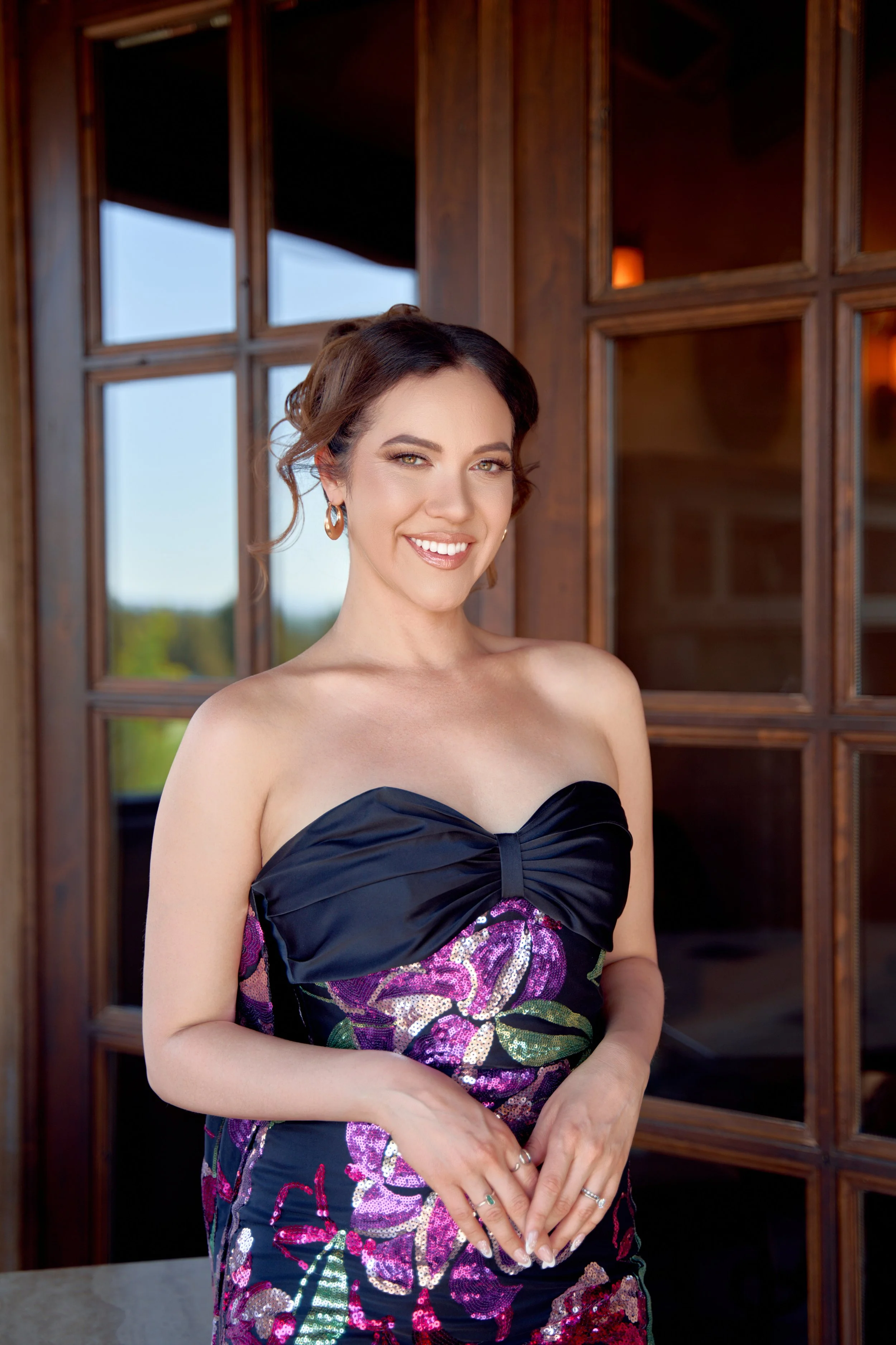 A woman wearing a strapless black dress with colorful floral patterns, standing indoors in front of a wooden-framed glass window, smiling.