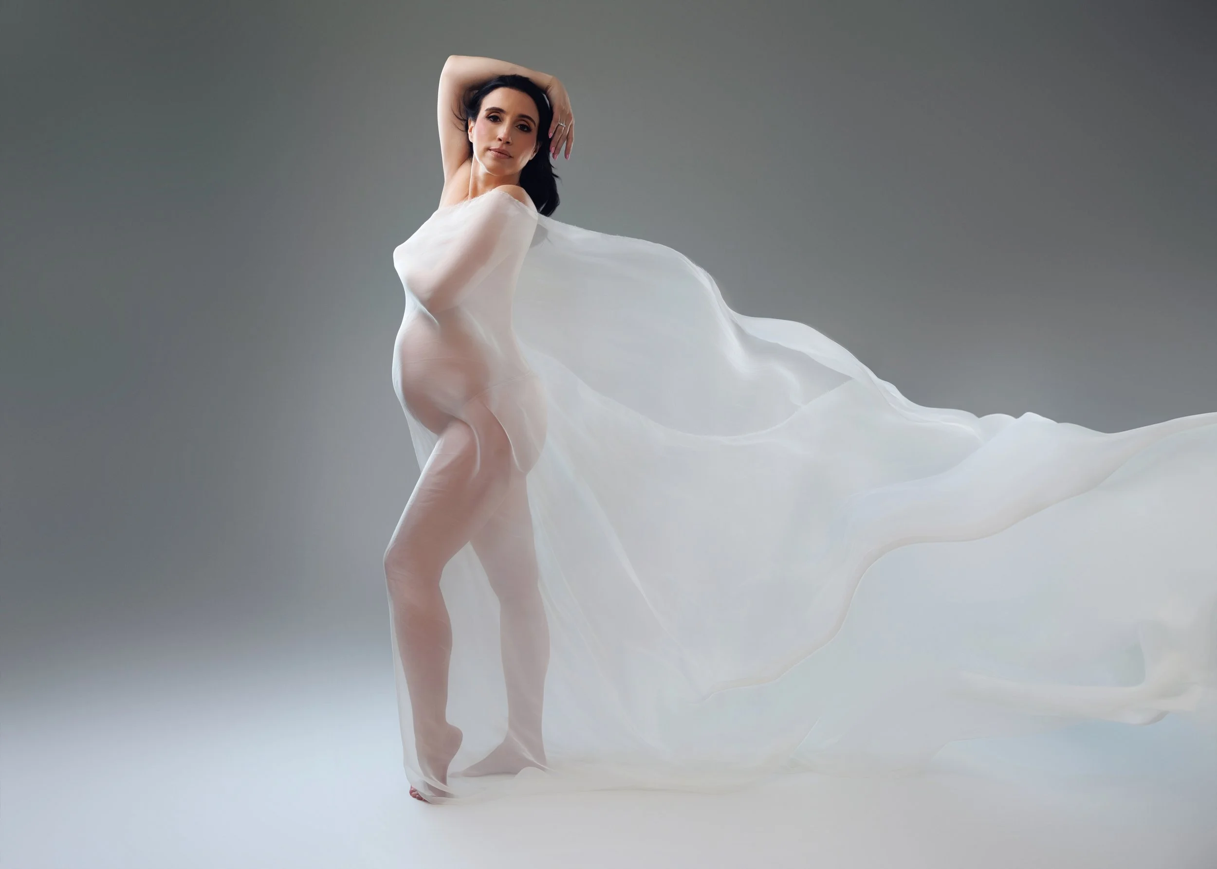 A woman with dark hair and makeup posing with white sheer fabric flowing behind her, standing on a white surface against a gray background.