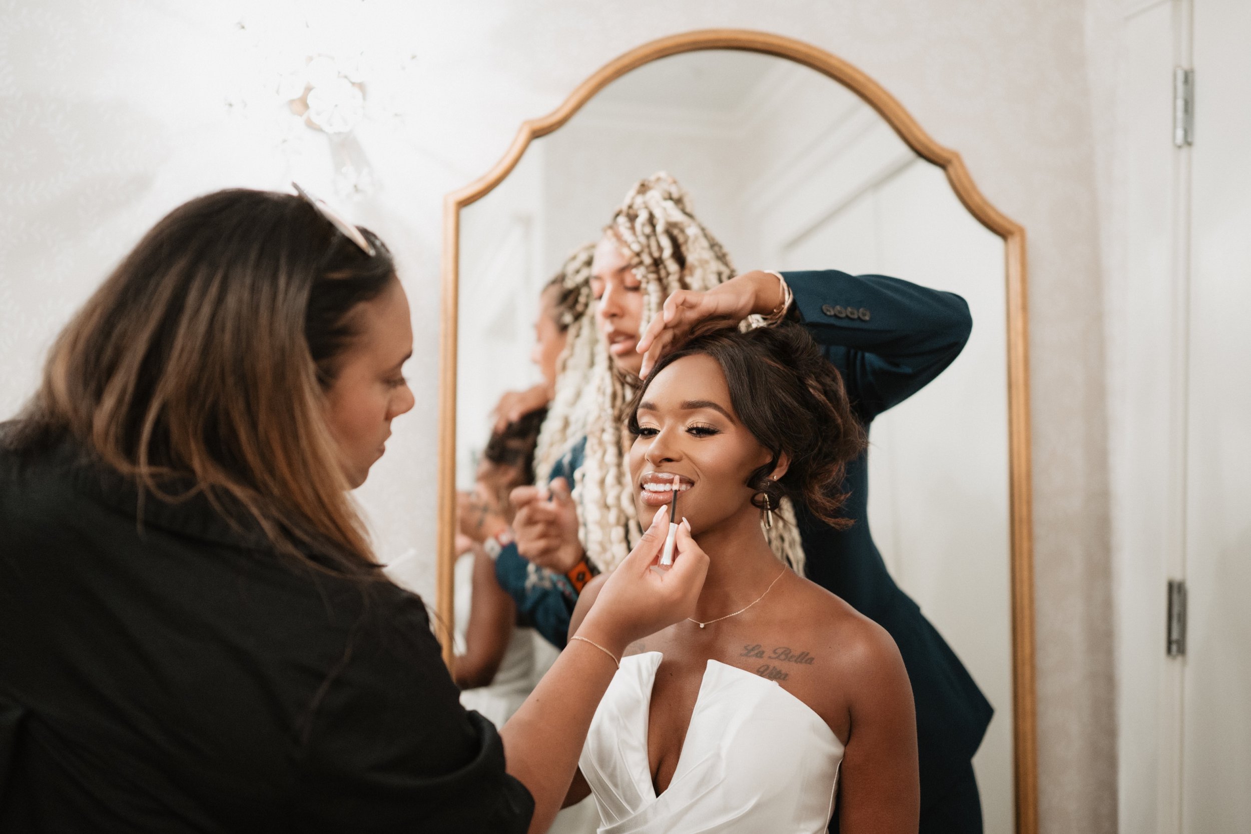 Makeup artist applying lipstick to a bride with a tattoo on her shoulder, while another woman styles the bride's hair in front of a mirror.