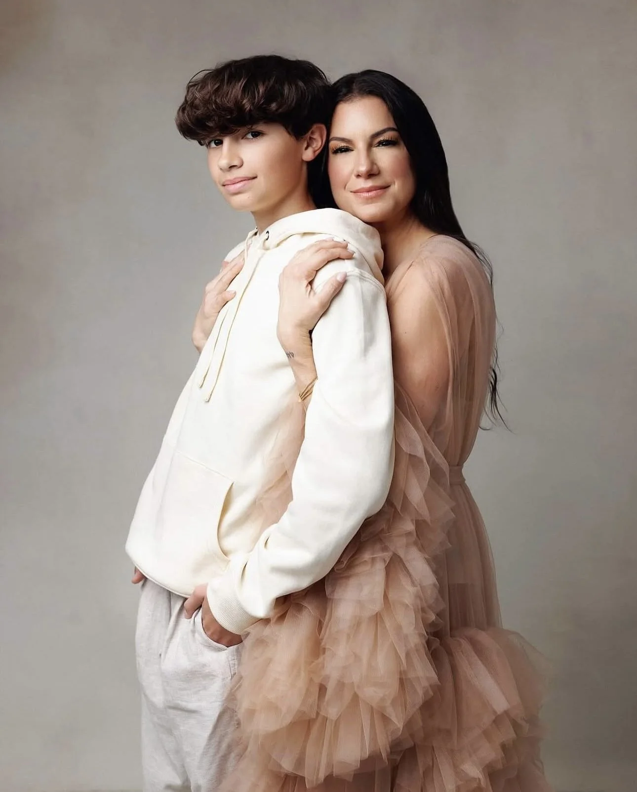 A woman and a teenage boy pose closely together, with the woman embracing the boy from behind. The woman has long dark hair and is wearing a peach-colored, ruffled dress. The boy has short, dark, wavy hair and is dressed in a light-colored hoodie and