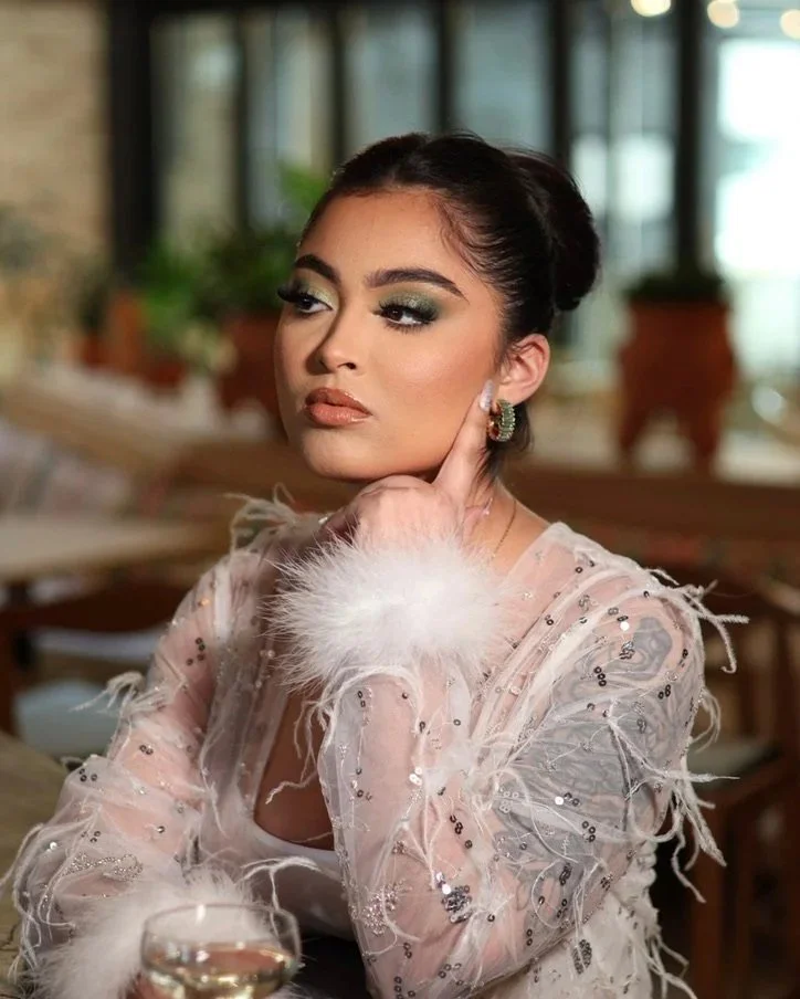 A woman with dark hair styled in an updo, wearing glamorous makeup with green eyeshadow, sitting at a table in a warmly lit restaurant or cafe. She is dressed in a light pink, sheer, sequin-embellished dress with feather accents on the sleeves, and is posed with her hand touching her face, looking to the side.