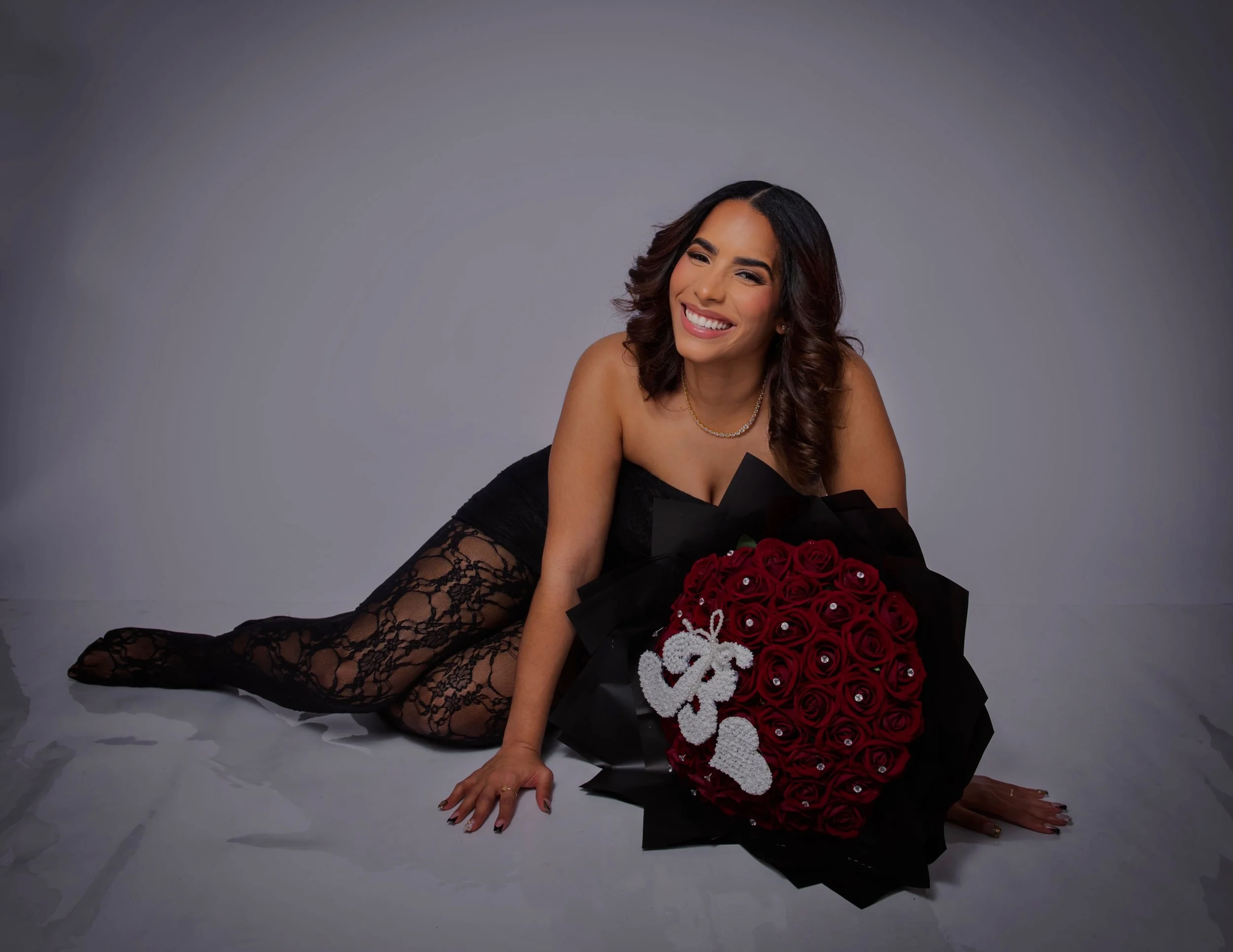 A woman in black lace stockings and a black dress is lying on the floor, holding a large bouquet of red roses decorated with pearls and a white bow, smiling at the camera.