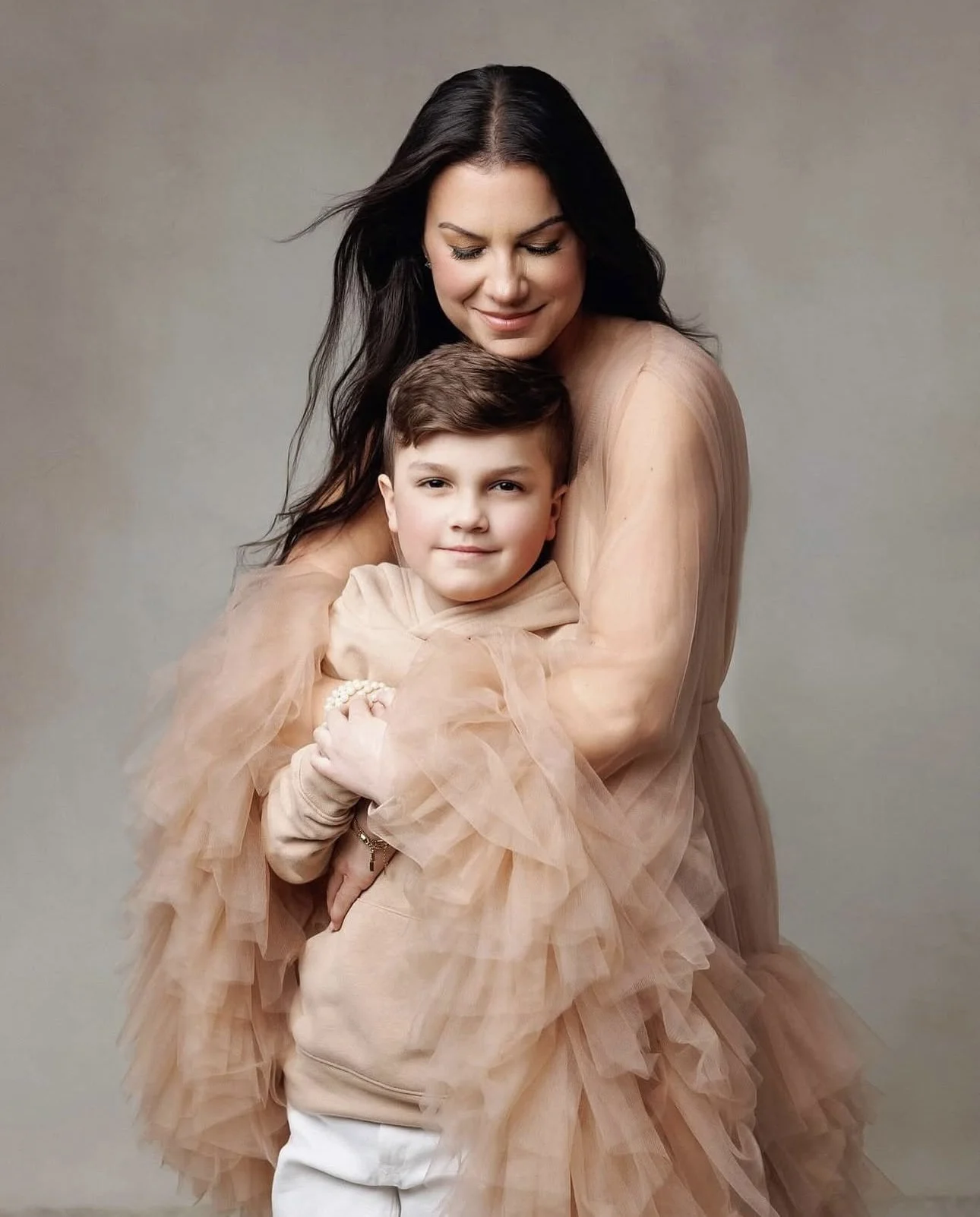 A woman and a young boy are hugging. The woman has long dark hair and is wearing a beige, ruffled dress. The boy has short brown hair and is wearing a beige hoodie and white pants. They are smiling softly against a neutral gray background.