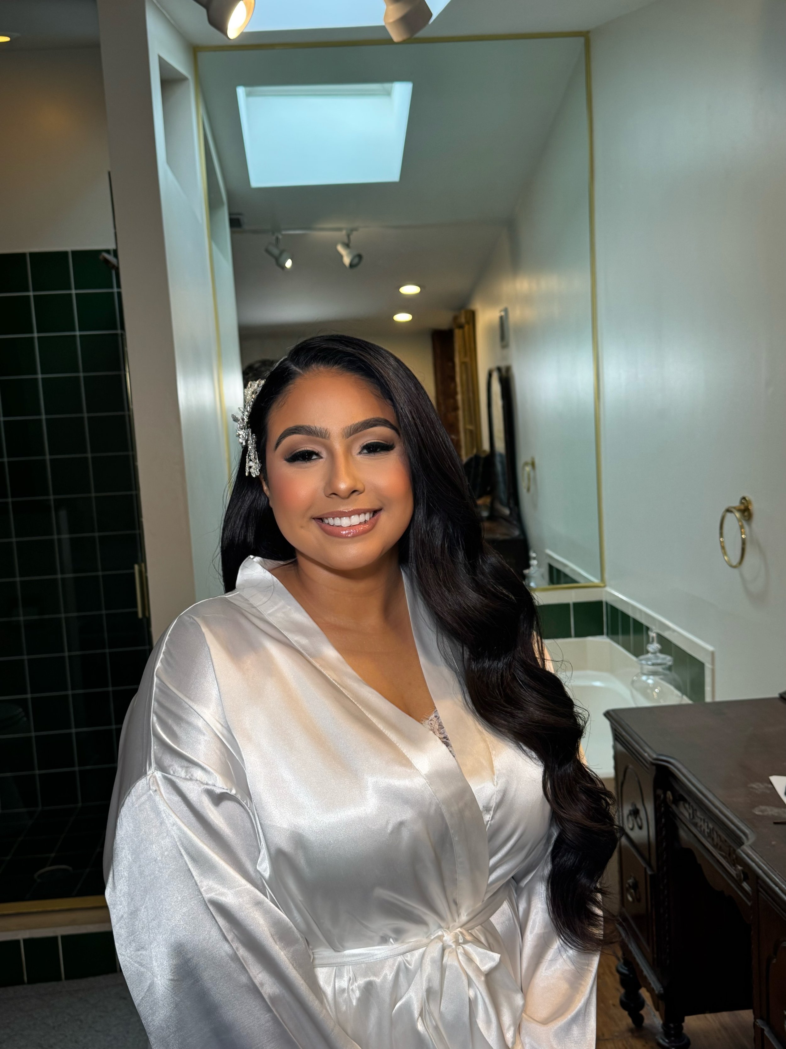 A woman with long black hair styled in loose waves, wearing a satin satin white robe, and a hair accessory, smiling in a mirror reflection in a bathroom.