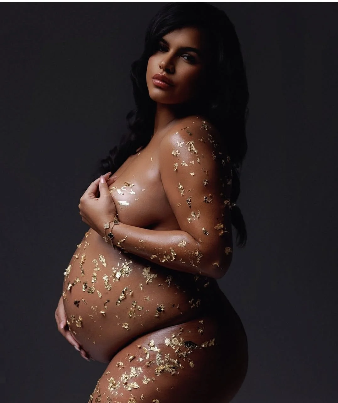 A pregnant woman with long dark hair and medium skin tone, covered in gold foil flakes, posing against a dark background.