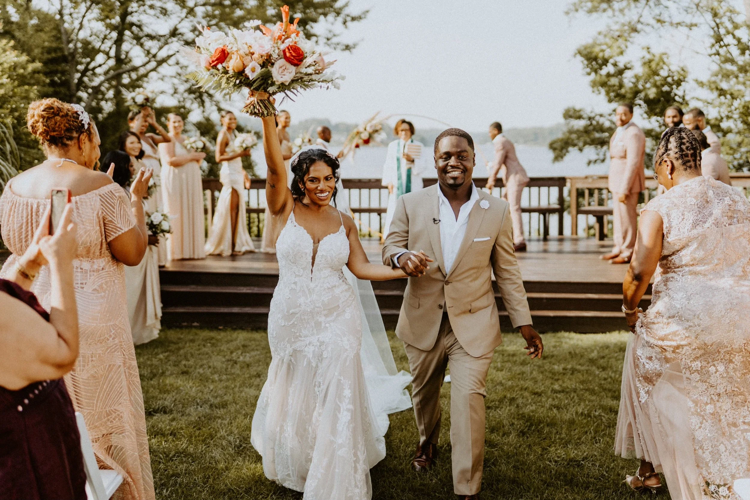 A newlywed couple holding hands and walking on grass during their outdoor wedding celebration, with the bride holding a large bouquet in the air, surrounded by friends and family, with a scenic water body and trees in the background.