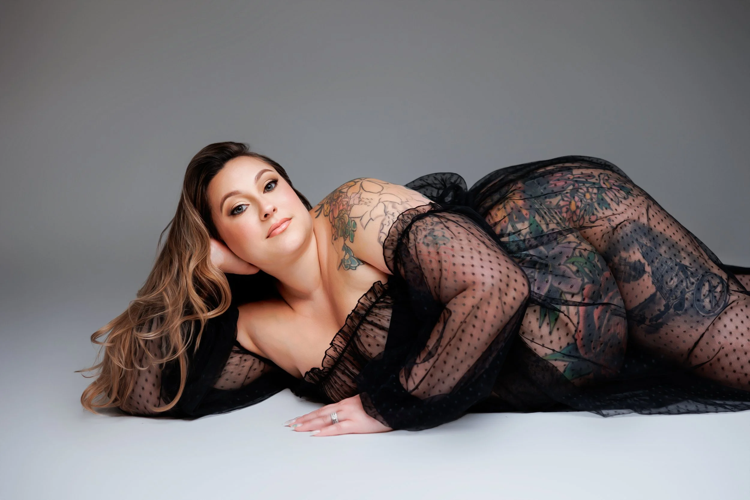 A Pregnant Woman with long hair lying on her side, wearing a sheer black polka-dot dress, showing colorful tattoos on her shoulder and arm, with a neutral gray background.