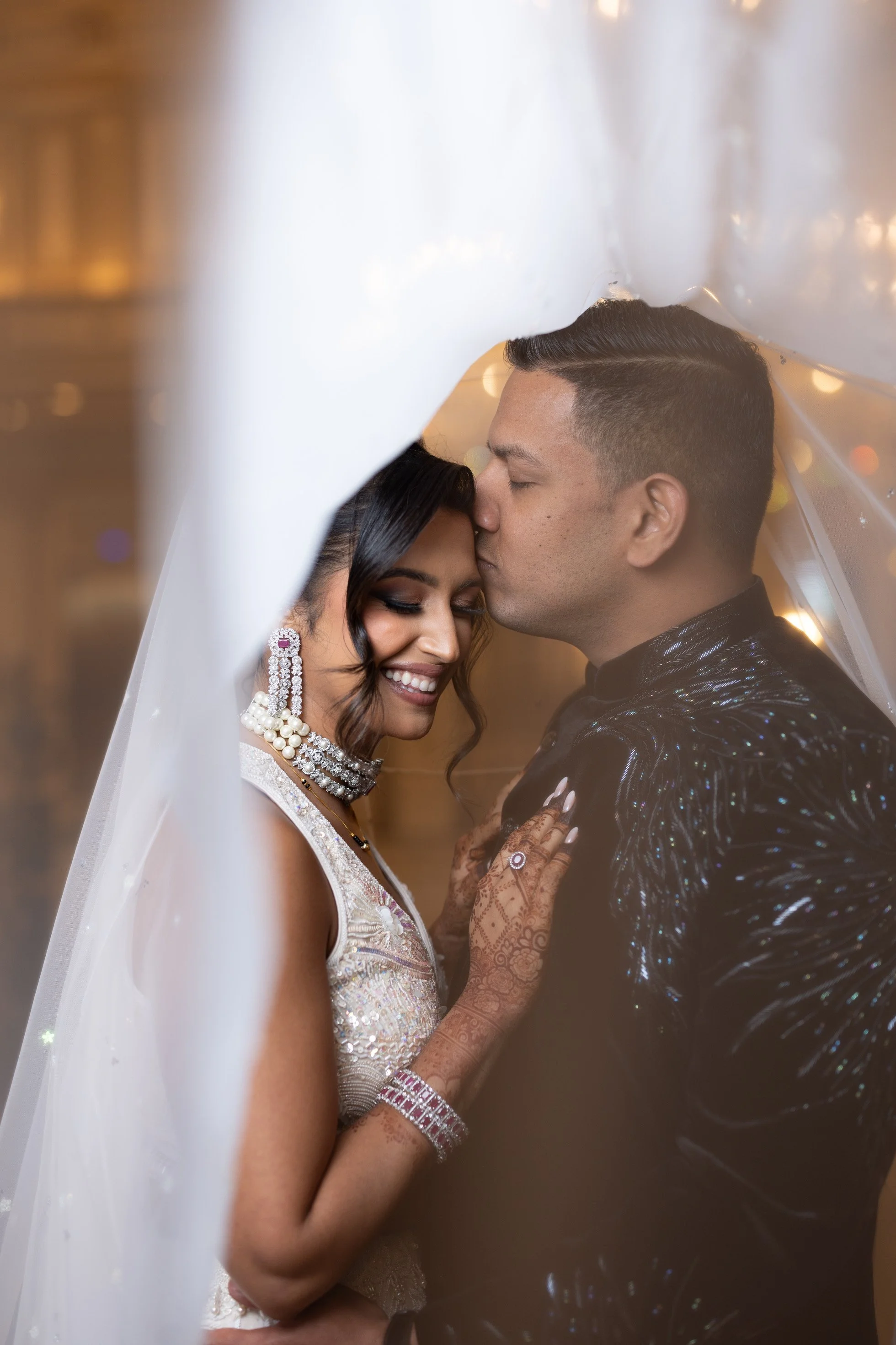 A newlywed couple shares an intimate moment under a white veil, with the groom kissing the bride's forehead. The bride is smiling, dressed in a traditional outfit with intricate jewelry and henna decorations on her hand, while the groom wears a black