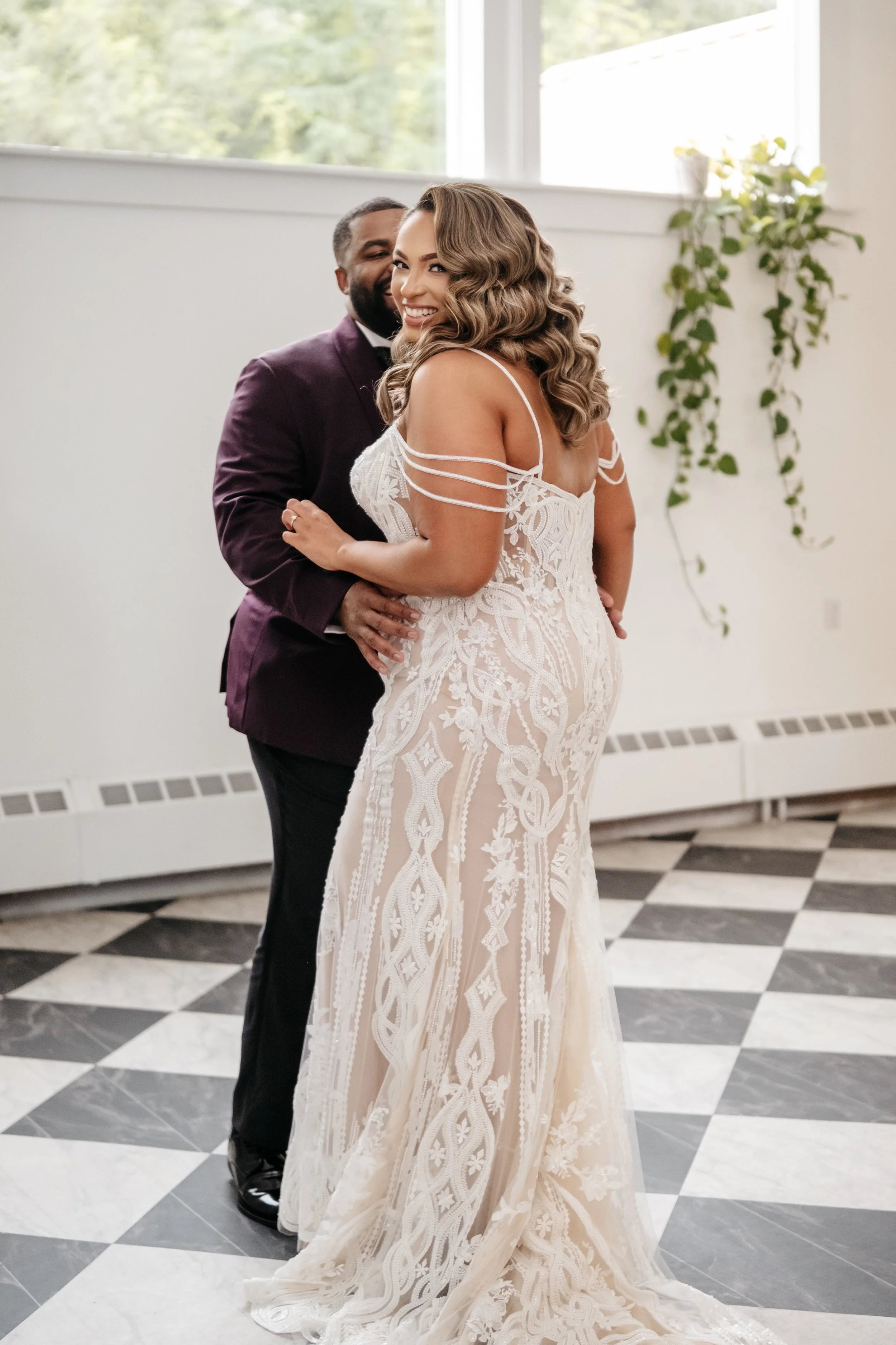A woman in a wedding dress and a man in a suit embrace indoors, smiling, with a checkered floor and windows in the background.