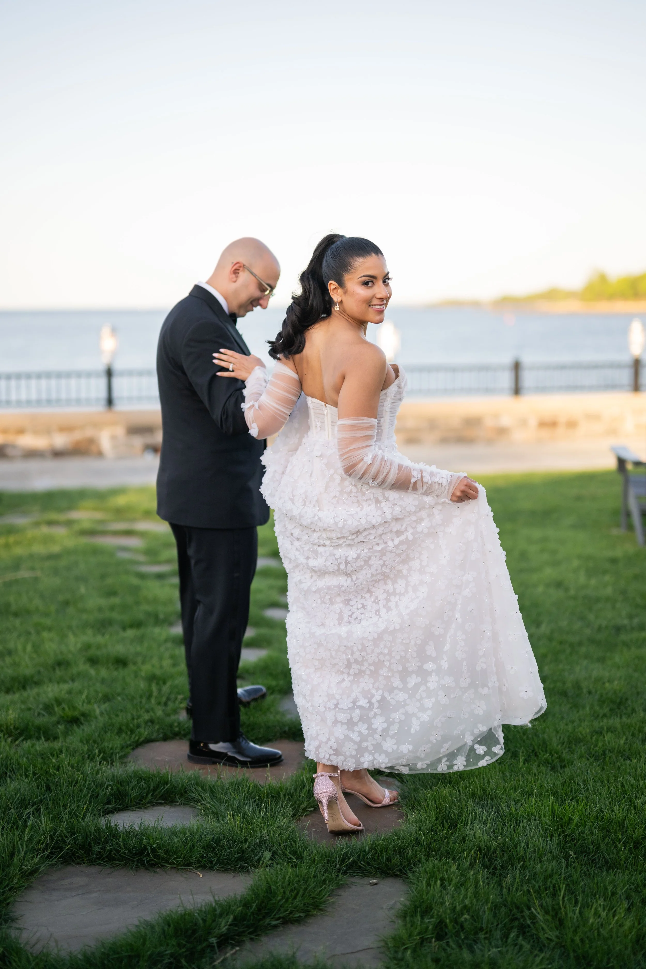 A bride in a white wedding dress with floral details, holding her dress and smiling, standing on a stone path on a grassy area near a body of water, with a man in a black suit behind her, outdoors during daylight.