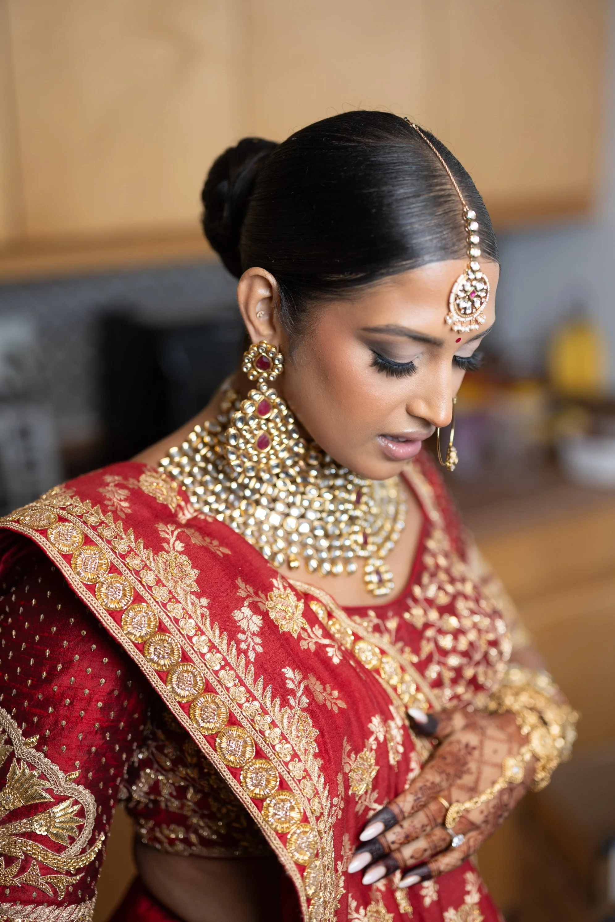 A woman dressed in traditional Indian bridal attire, with elaborate jewelry including earrings, nose ring, necklace, and head jewelry, is adjusting her outfit and looking down.