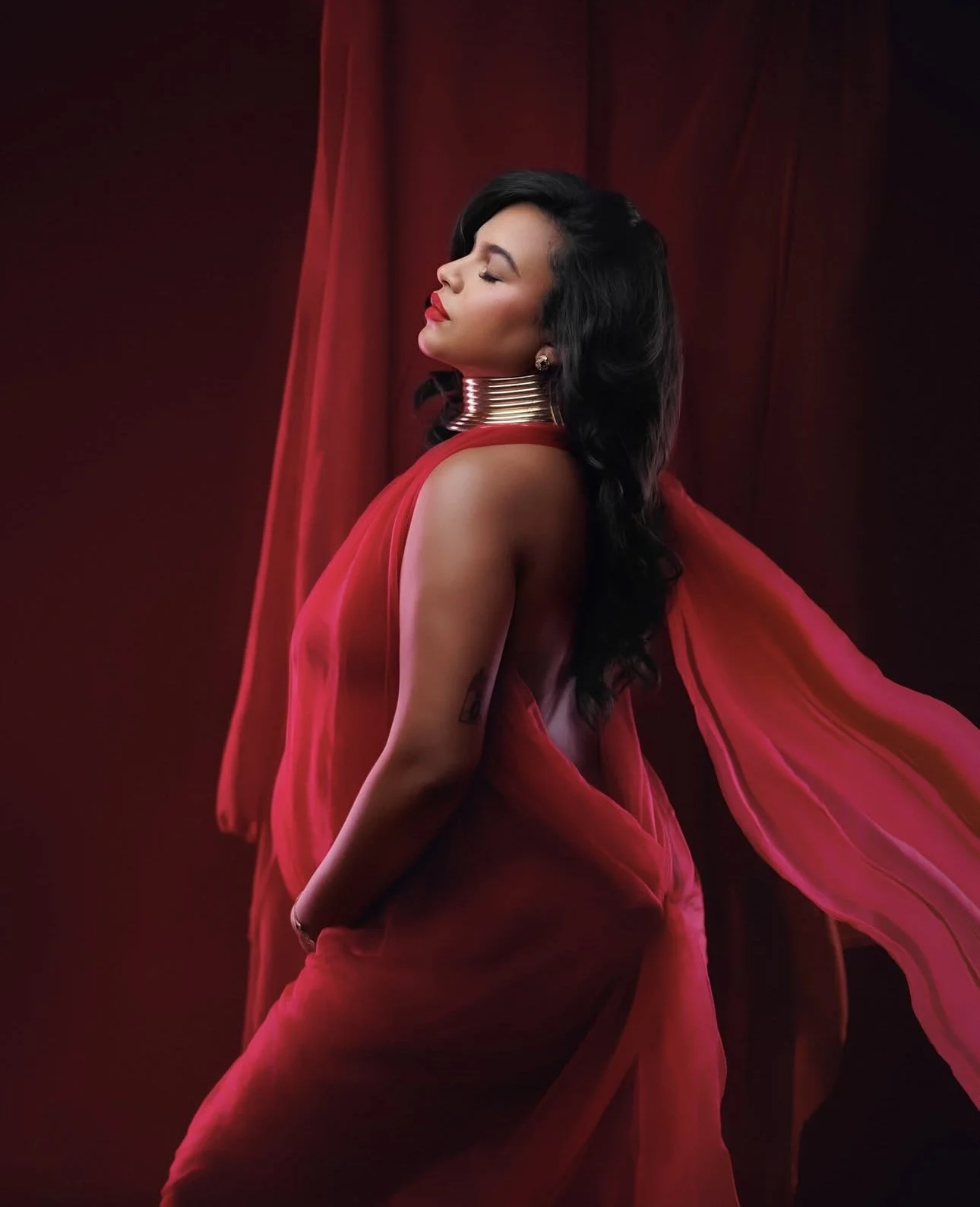 Woman with closed eyes wearing a flowing red dress, large metallic neck ring, and earrings, posing against a red background with curtains.