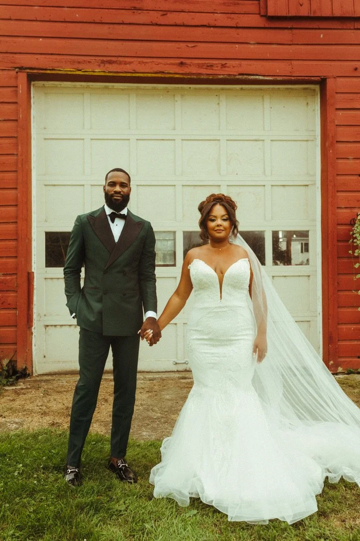 Bride and groom in wedding attire holding hands outside in front of a white garage door with a red wooden building.