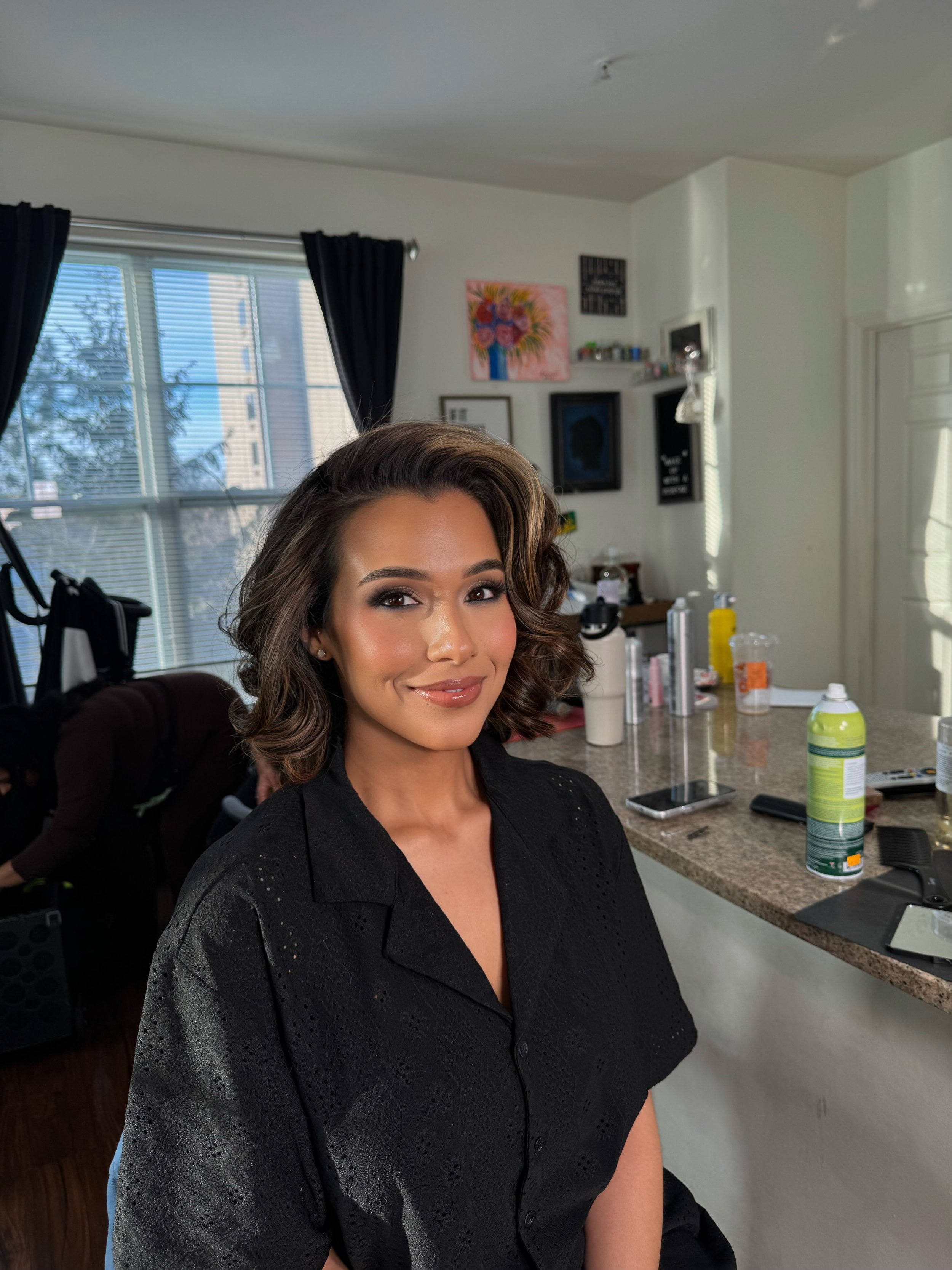 A woman with styled curly hair and makeup sits in a living room or kitchen area, smiling at the camera.
