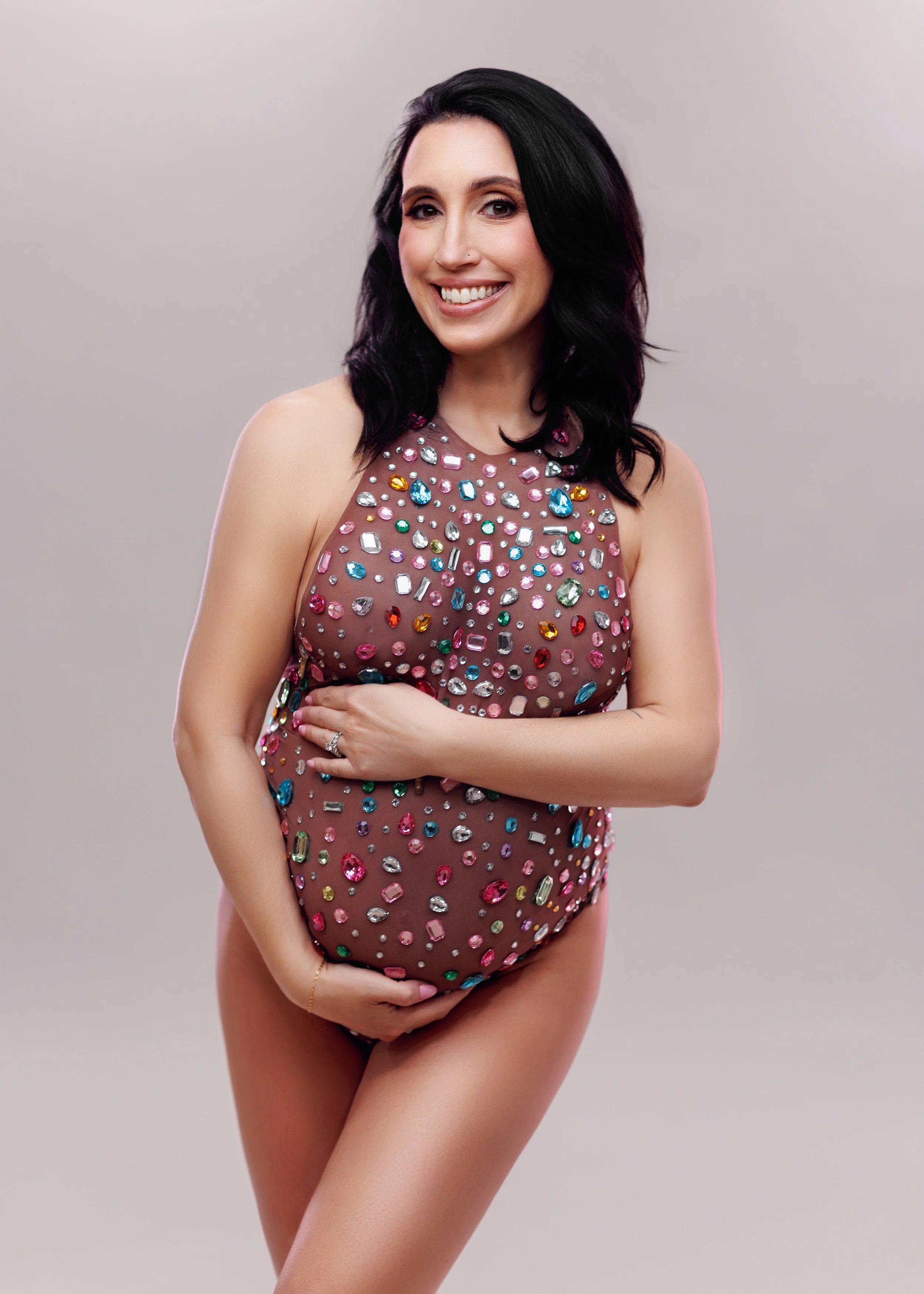 A smiling pregnant woman with black hair, wearing a sheer dress decorated with colorful gemstones, holding her belly and standing against a plain background.