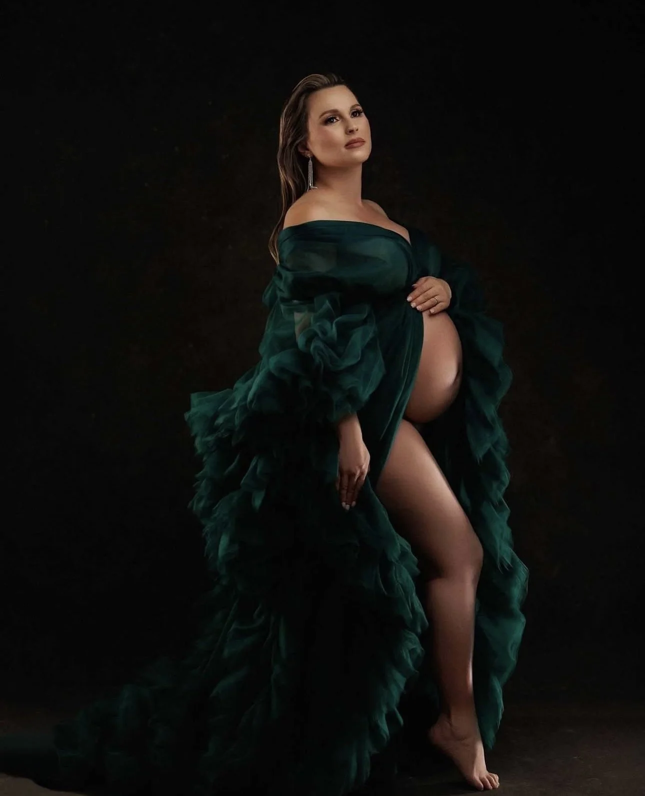 A pregnant woman in an elegant green gown with ruffled sleeves poses against a dark background.