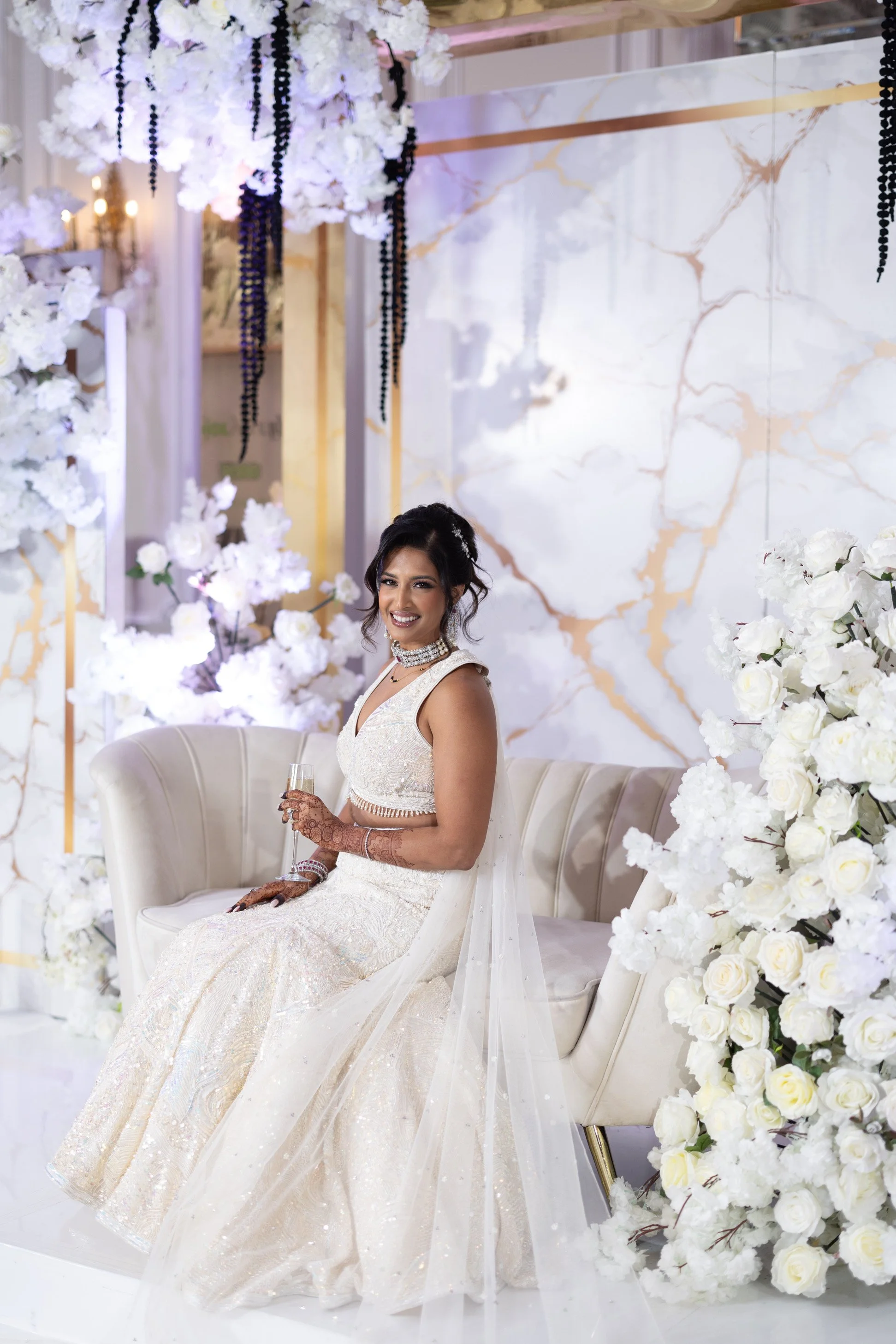 A bride in a white wedding gown and veil, sitting on a cream-colored sofa surrounded by white flowers and decorative elements, holding a glass of champagne and smiling.