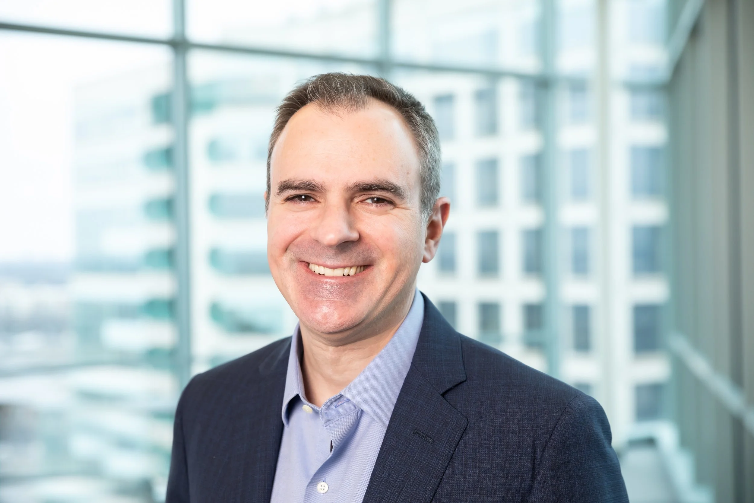Headshot of Ralph DeBerardinis, he is smiling and standing in front of a series of windows.