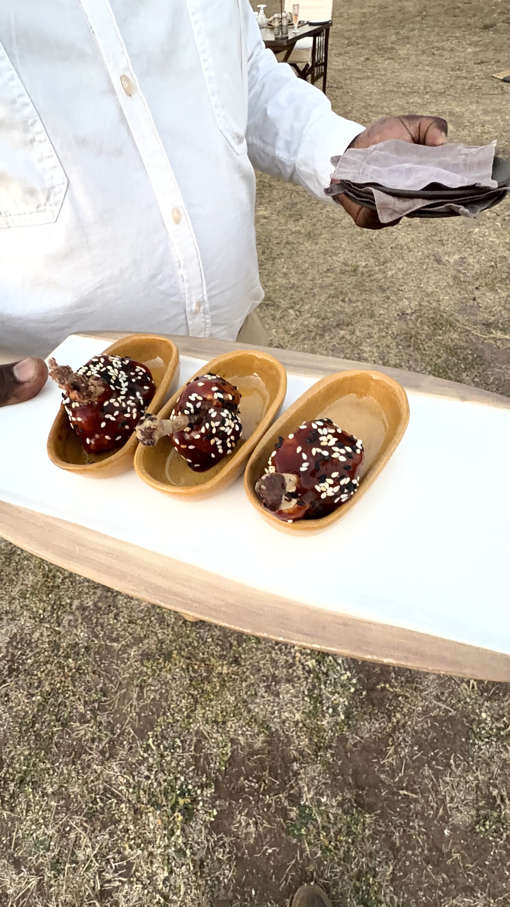 A member of the Mila team serving sesame-glazed snacks on a wooden tray outdoors in the evening