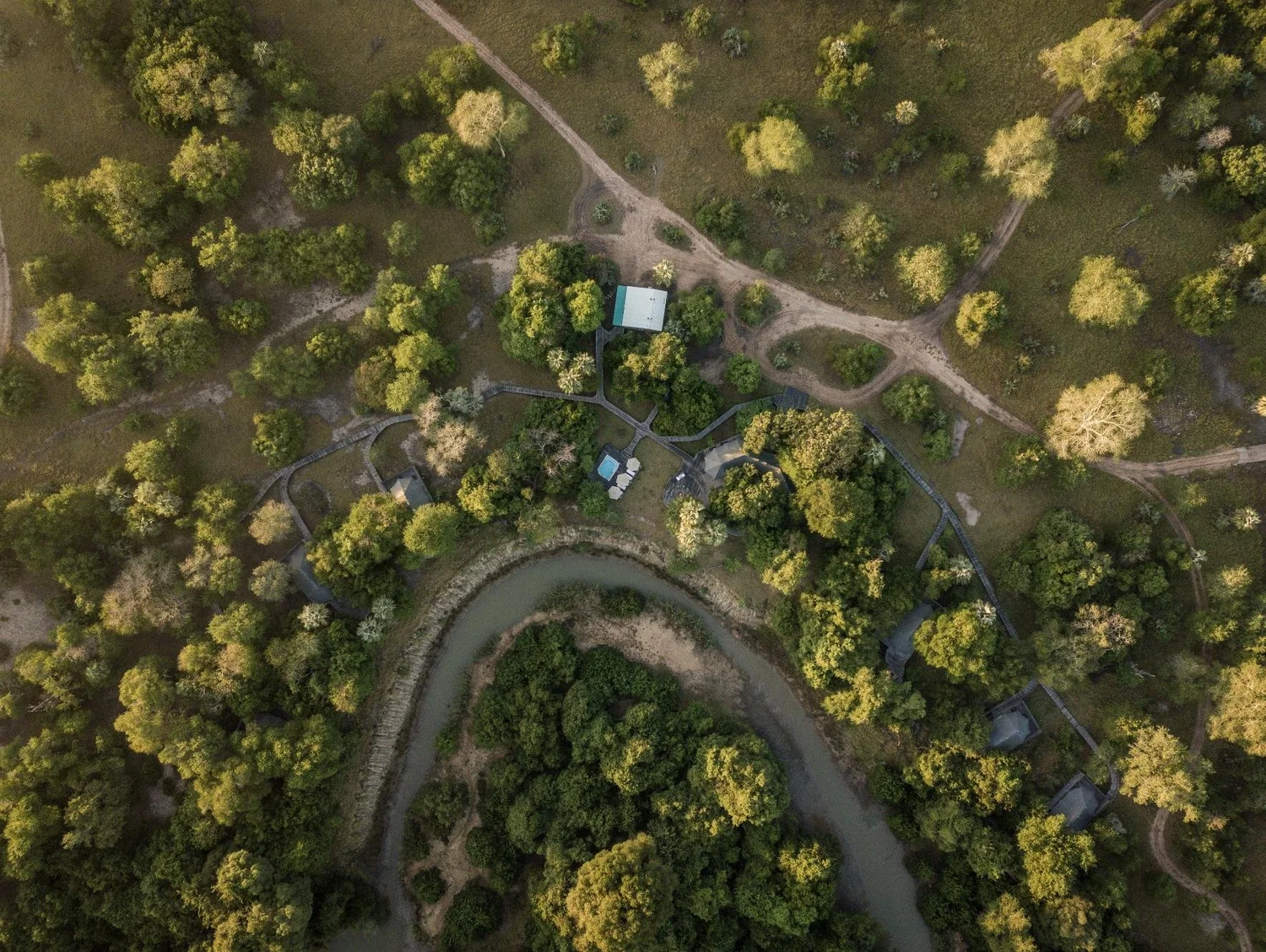 An aerial view of a winding river surrounded by lush green trees, with some pathways and small buildings near the river and in the forested area.