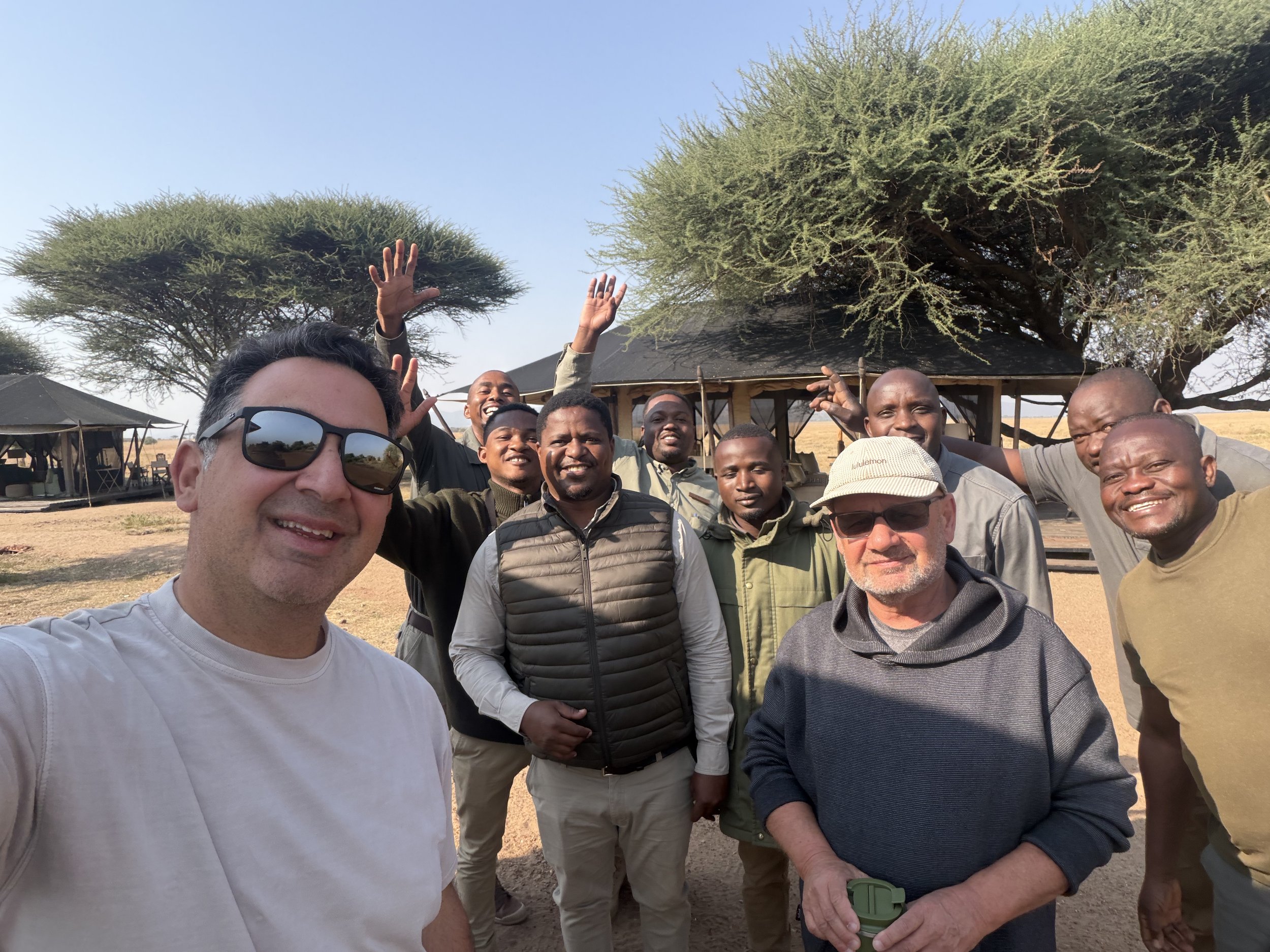 Group selfie with members of the Mila camp team, several waving, in front of the camp's tents and acacia trees