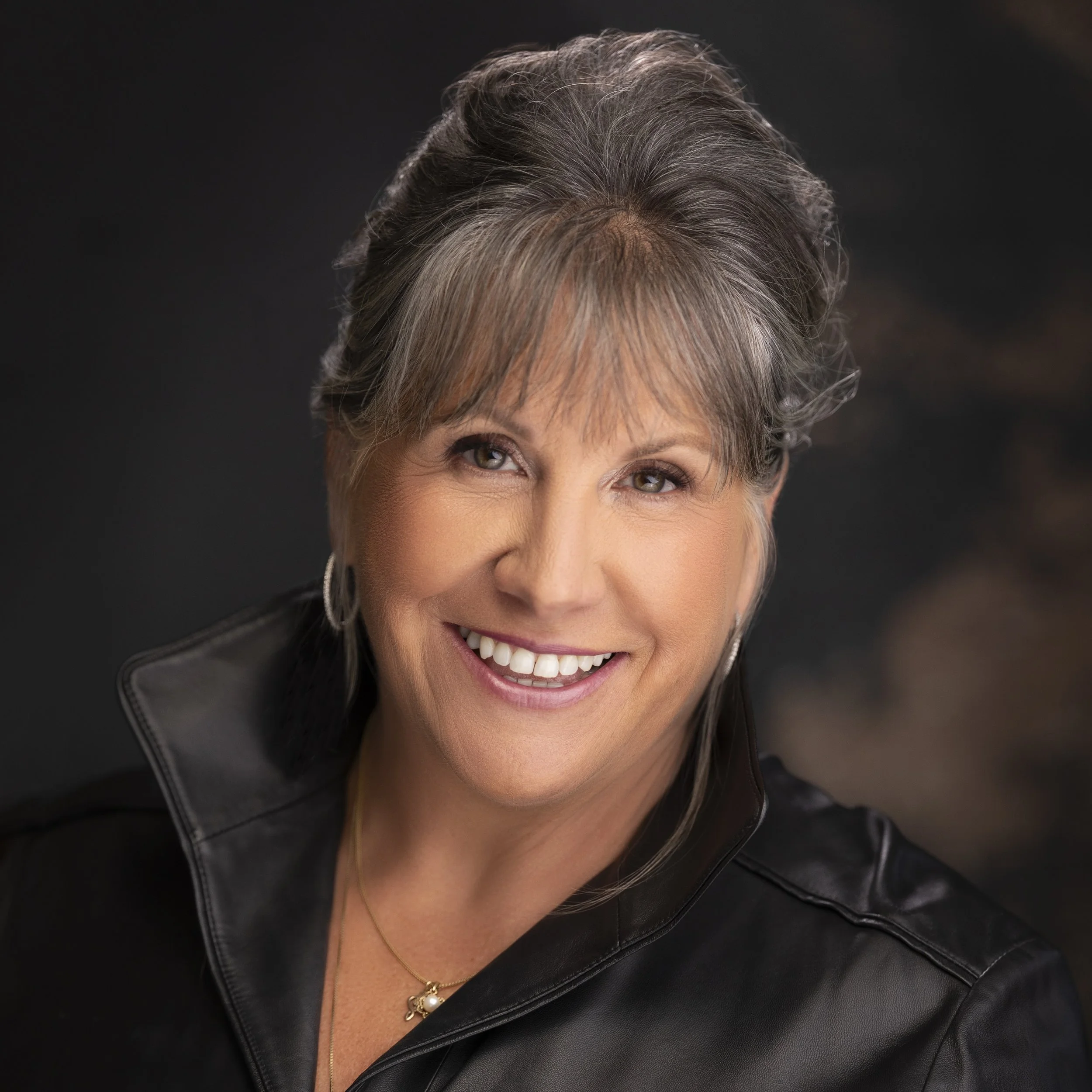 Close-up photo of a woman with short, tousled reddish hair, wearing a black top, smiling and resting her chin on her hand.