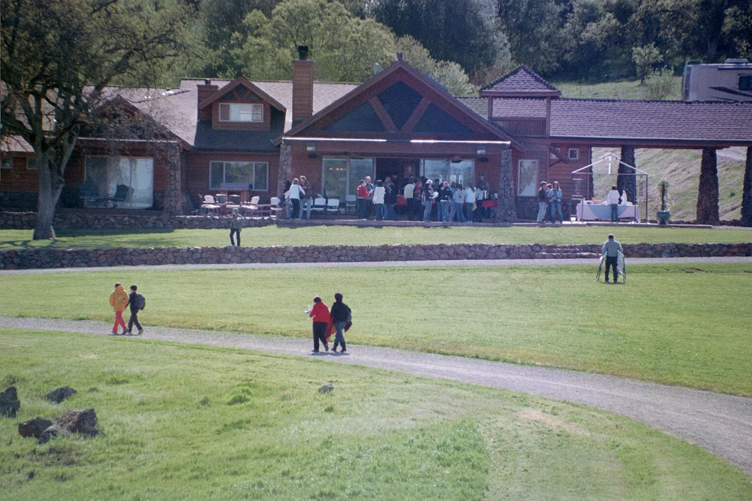 A large group of people gathered outside the main community house, situated on a grassy area with trees in the background. Some individuals are walking on a gravel path in the foreground.