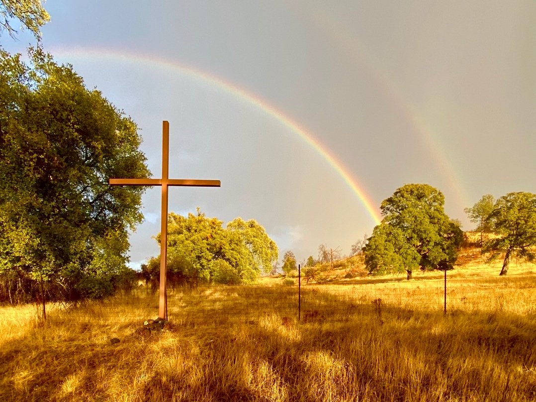 A large metal cross standing in a grassy field with trees, a double rainbow in the sky, and a fence in the distance.