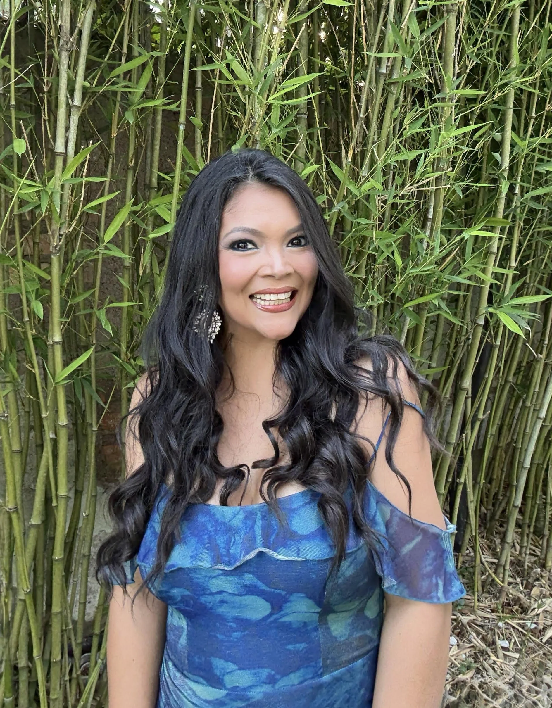 Karen Zeiter with long black curly hair, wearing makeup, jewelry, and a blue dress, smiling, standing in front of a bamboo plant.