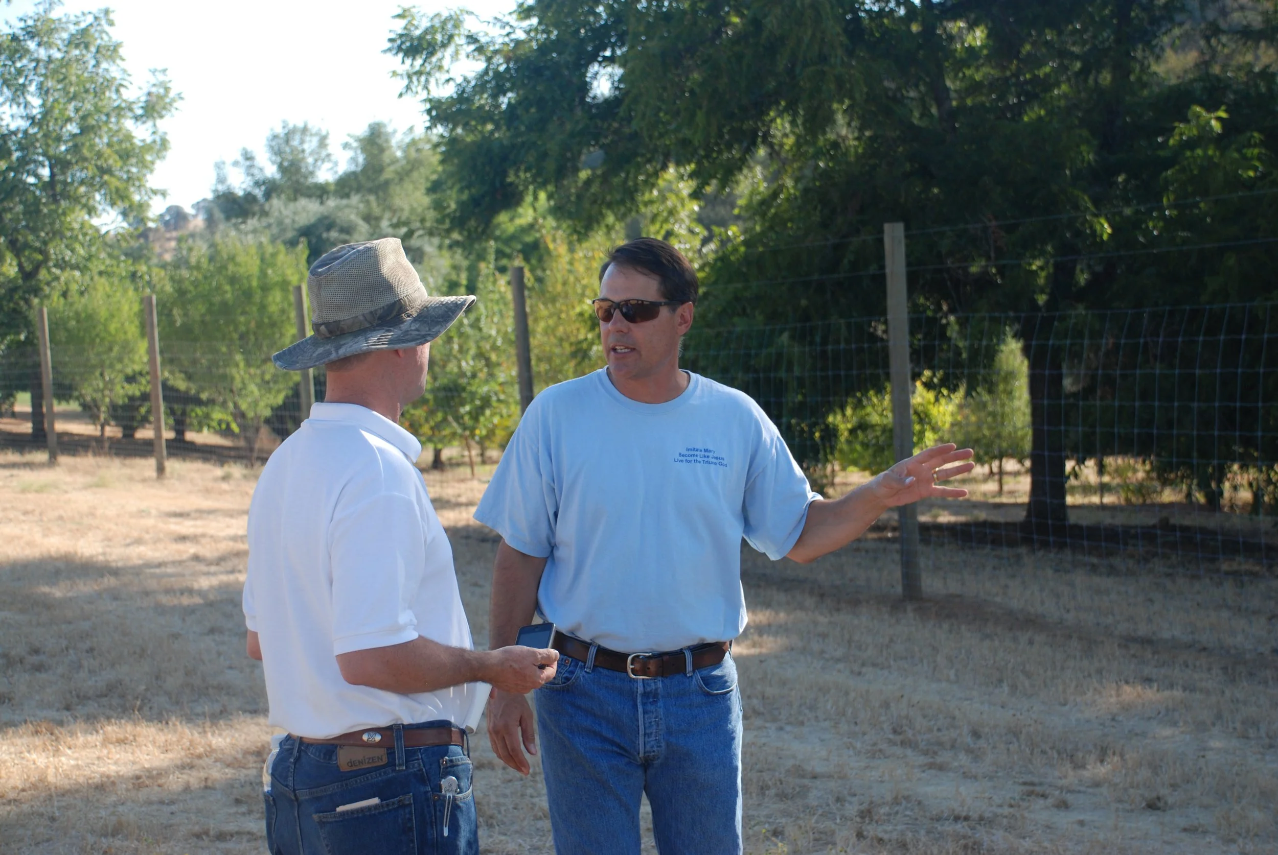 Two men having a conversation outdoors in a rural setting with trees and a wire fence in the background. One man is wearing a wide-brimmed hat and white shirt, and the other is wearing sunglasses and a light blue T-shirt.