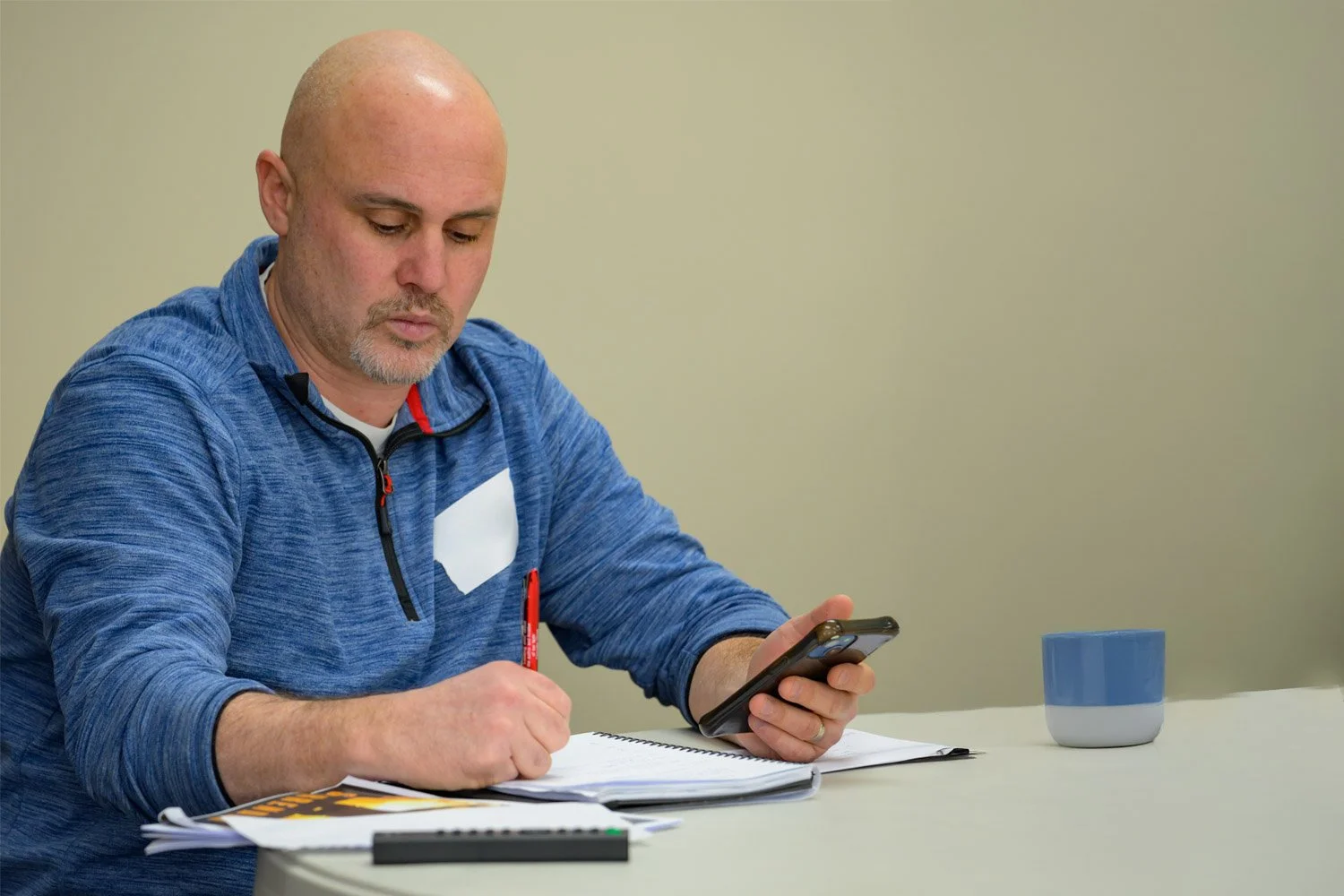 A man with gray hair and glasses, wearing a quilted jacket, is reading a Bible and taking notes with a pen in his right hand, seated in a crowd.