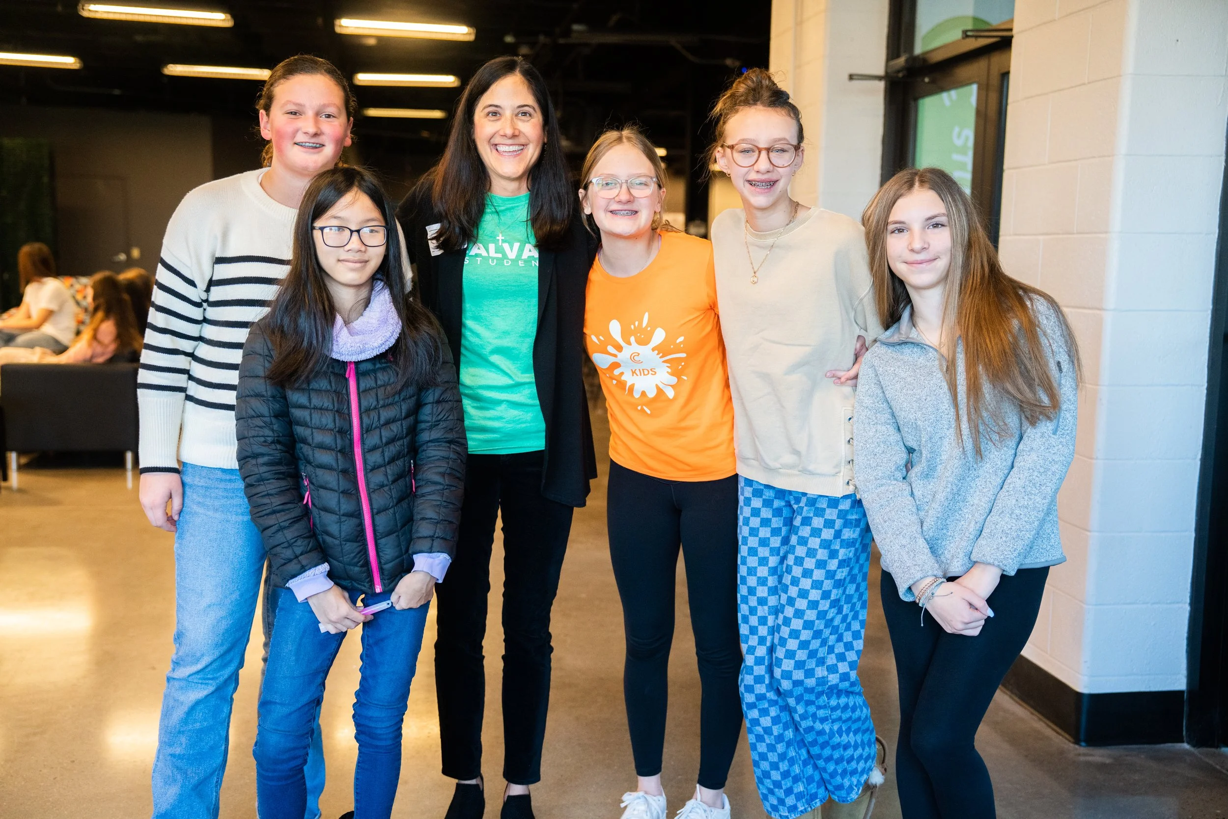Group of six girls and one woman standing indoors, smiling for the camera. The woman is in the center, wearing a green T-shirt with white text and a black jacket. The girls around her have casual clothing, with some wearing glasses and one girl with braces. The background shows other people sitting on a couch.