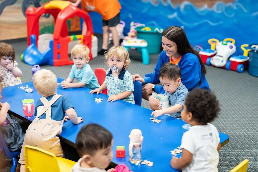 A group of young children and three adult women posing together in a colorful classroom, smiling and making playful gestures.