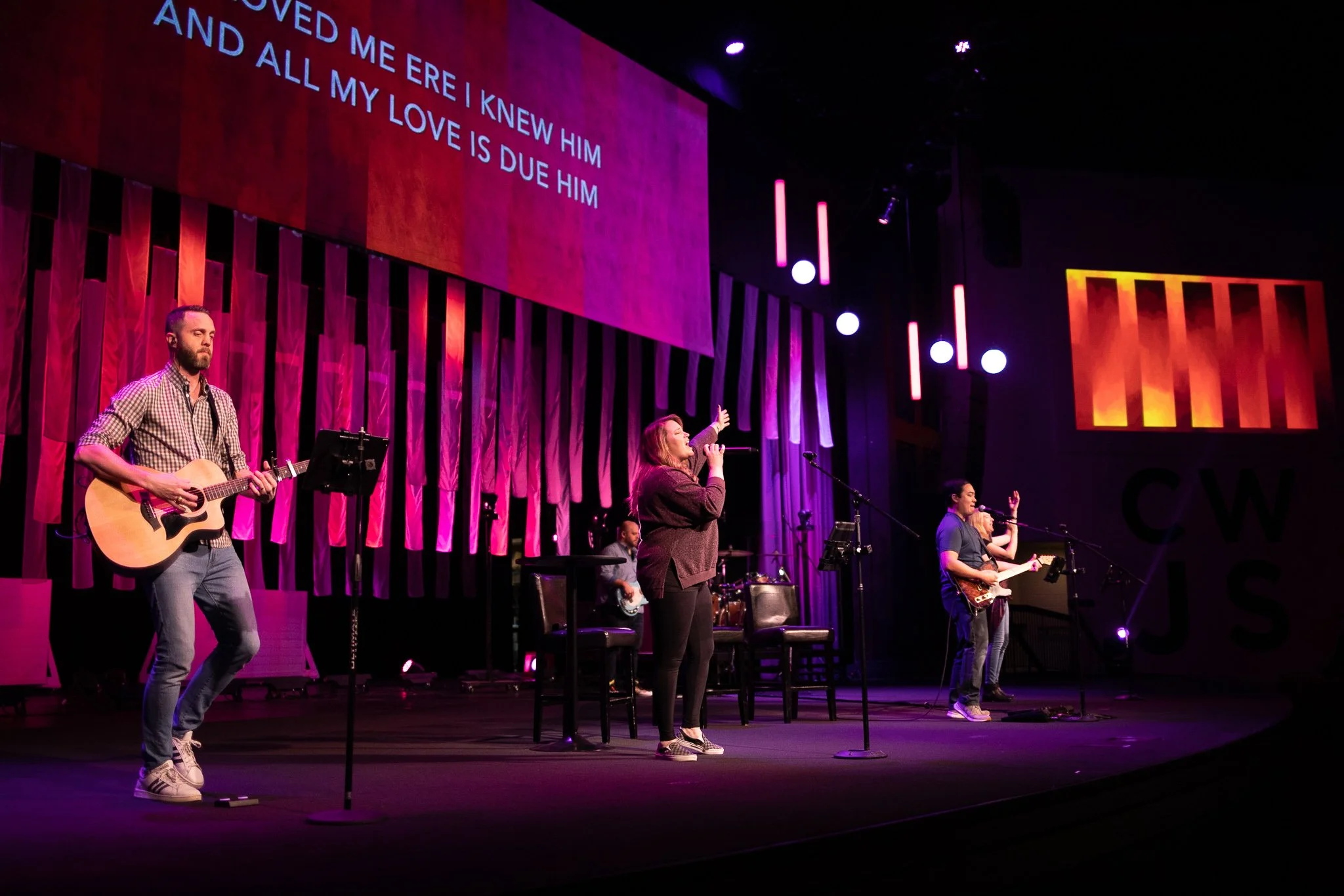 A band performing on stage with purple and pink lighting, including a guitarist, a singer, a bassist, and a drummer. The large screen behind displays lyrics: "LOVED ME ERE I KNEW HIM AND ALL MY LOVE IS DUE HIM."