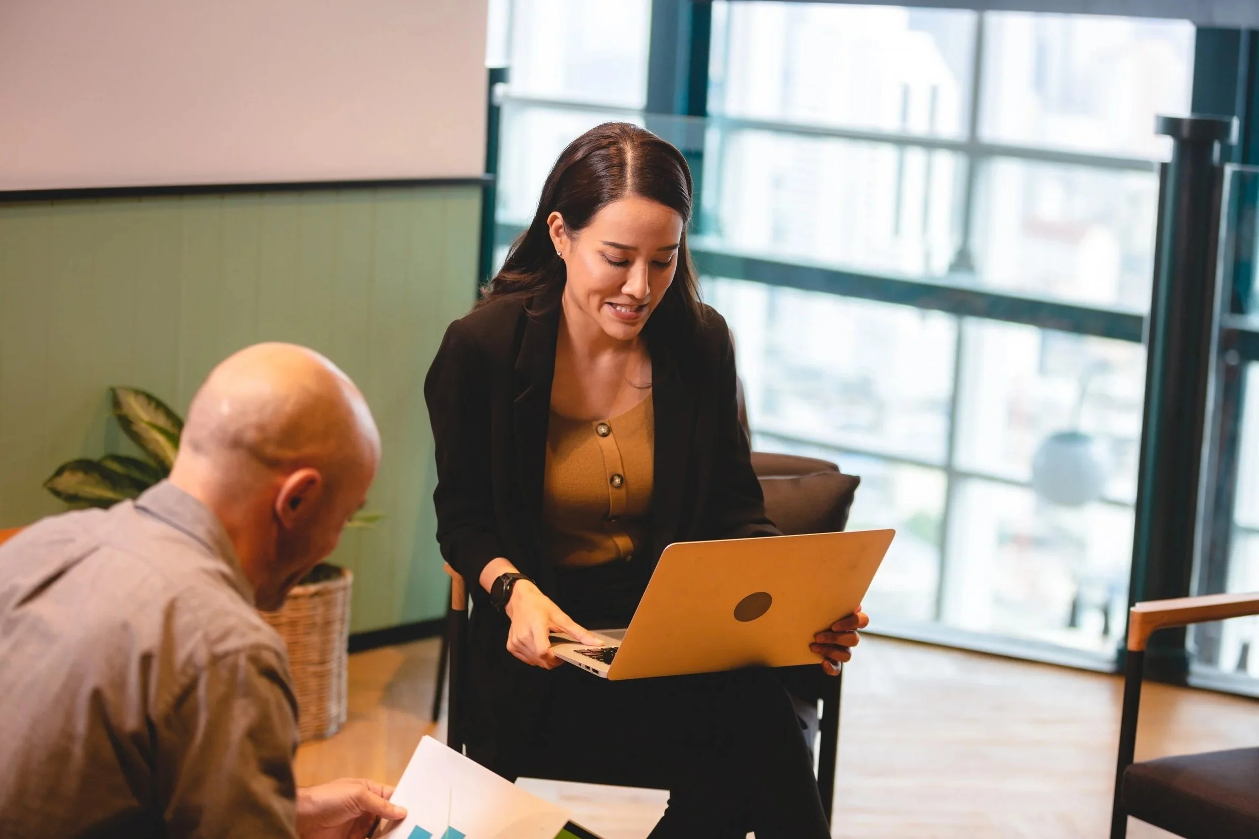 A woman showing a laptop to a man holding a paper in a modern office setting with large windows.