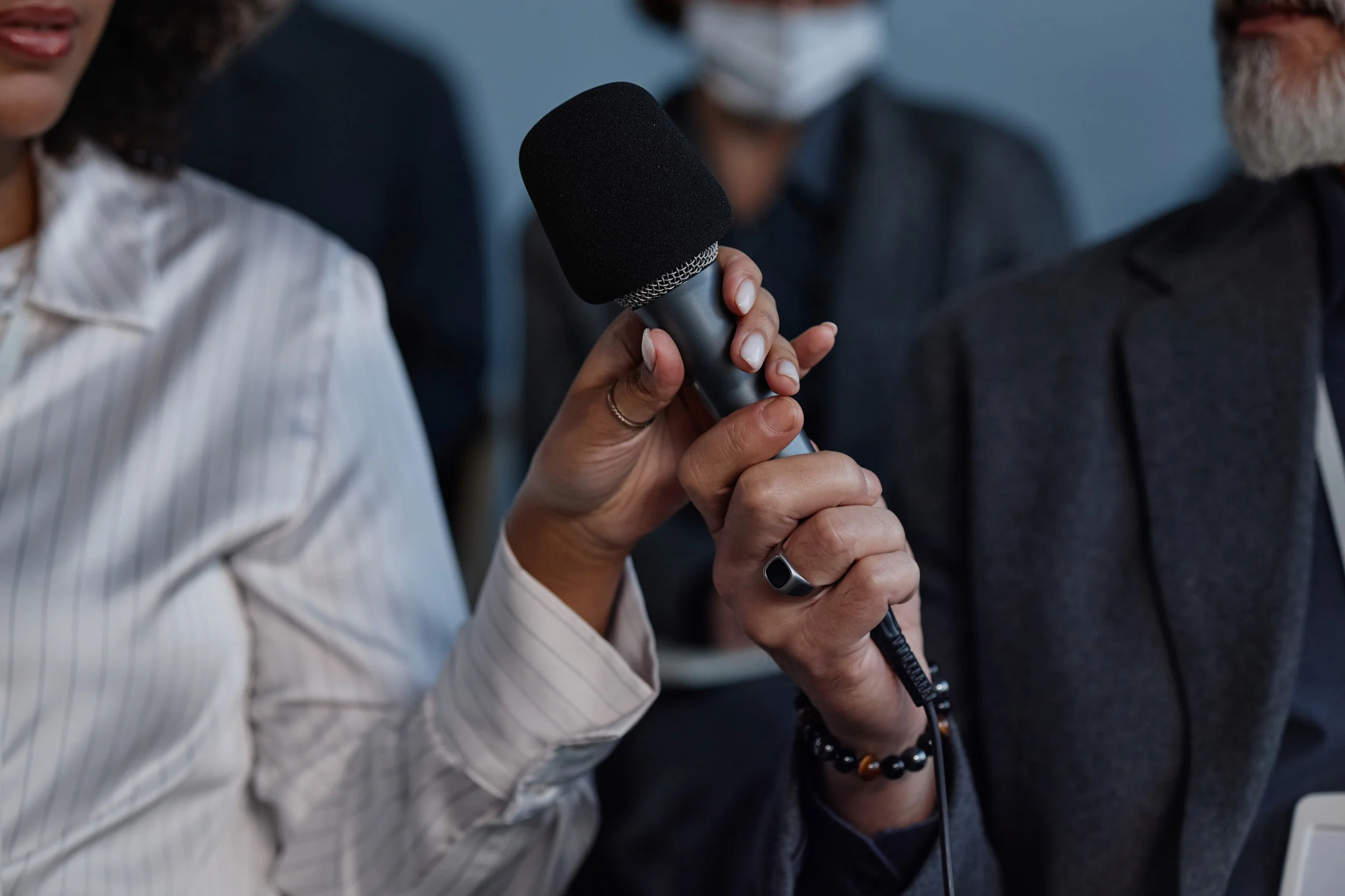 A woman holding a microphone during a group interview or press conference, with people blurred in the background.