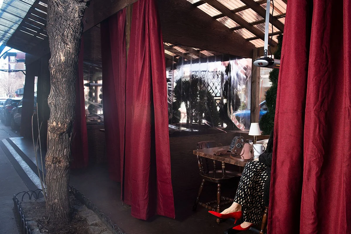 This is a color photograph, depicting an outside eating area with red curtains, behind which person is seated. The seated person cannot be seen, except for her legs and red high-heels. Photograph is by Nancy A. Scherl.