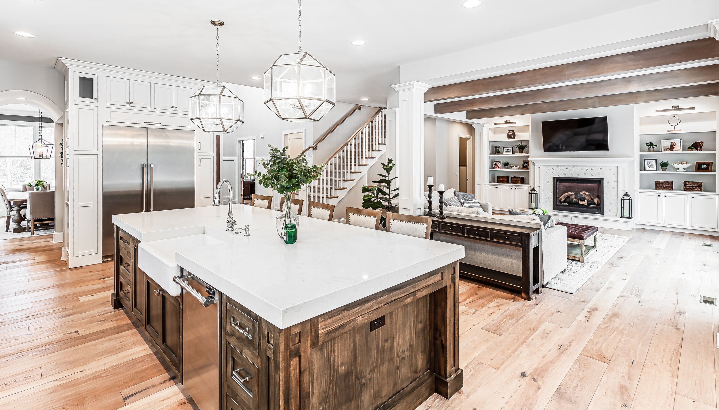 This warm and inviting open-plan living space includes a kitchen and living area. The kitchen features a large central island with dark wood and a white marble countertop. Above the island, geometric pendant lights add a contemporary flair. The view 