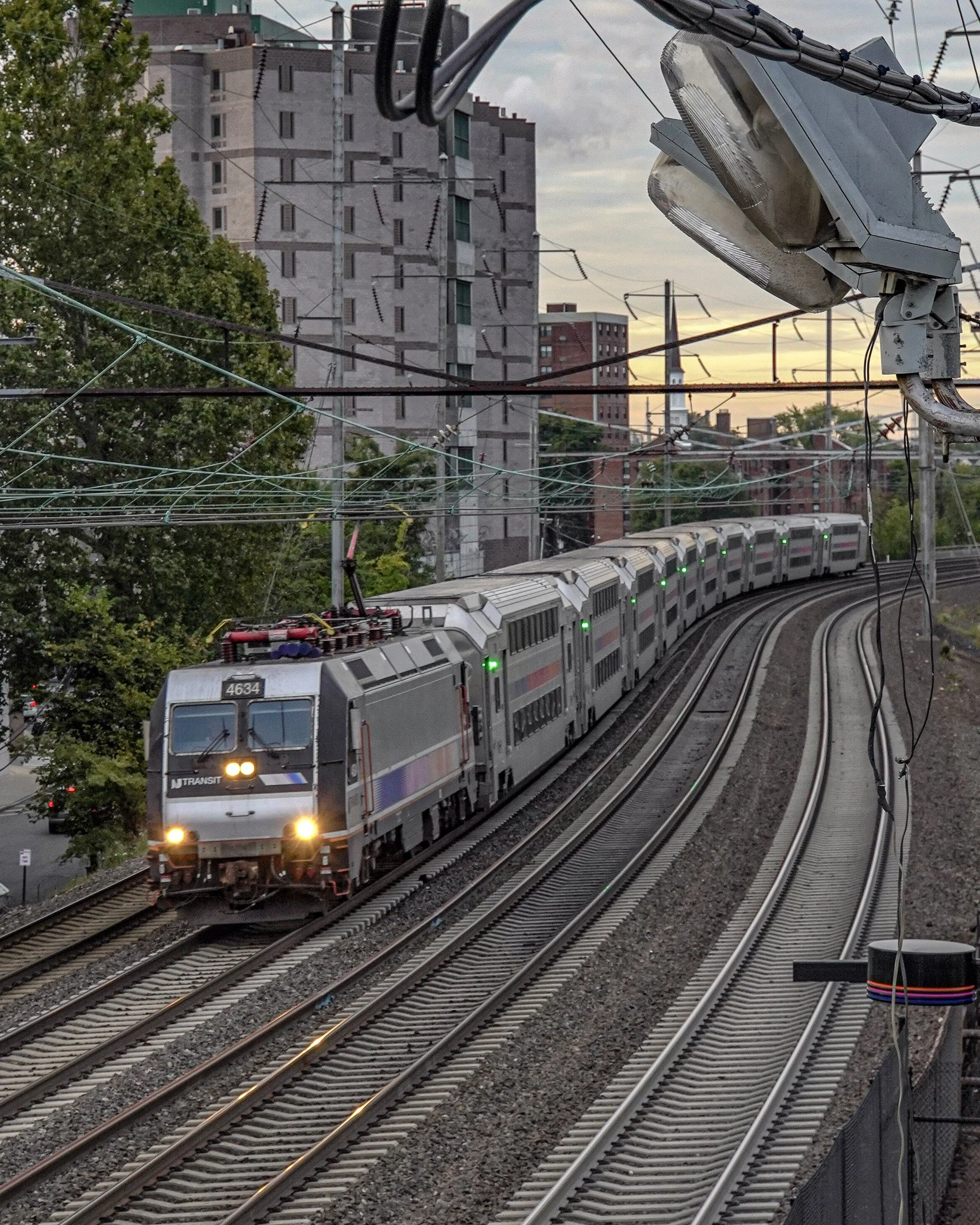 A commuter train traveling on elevated railway tracks during dusk, with overhead electrical wires and a cityscape with tall buildings in the background.