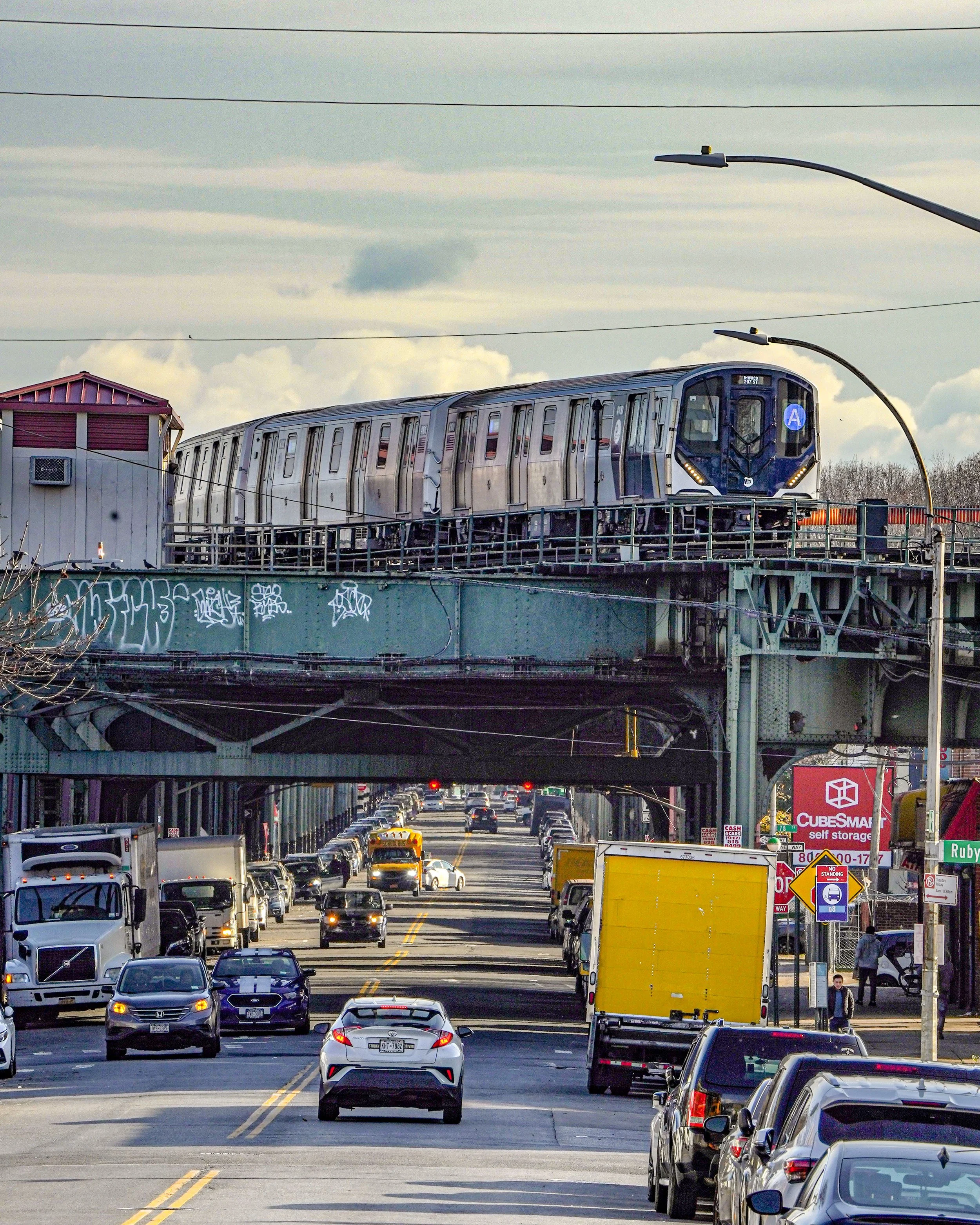 A subway train traveling above a busy street filled with cars, trucks, and pedestrians, with a cloudy sky in the background.