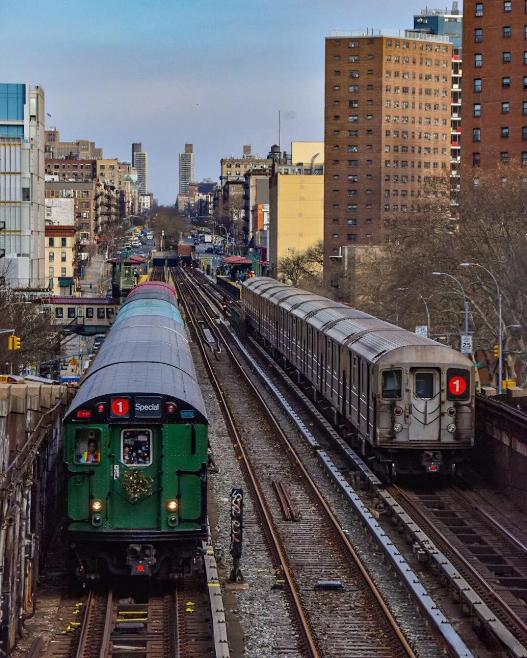 City scene with two subway trains on parallel tracks, one decorated with a wreath, and city buildings in the background.