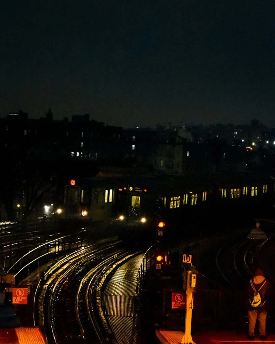 Nighttime view of a cityscape with a train on elevated tracks and a person waiting at a station, illuminated by station lights, with a dark sky and city buildings in the background.