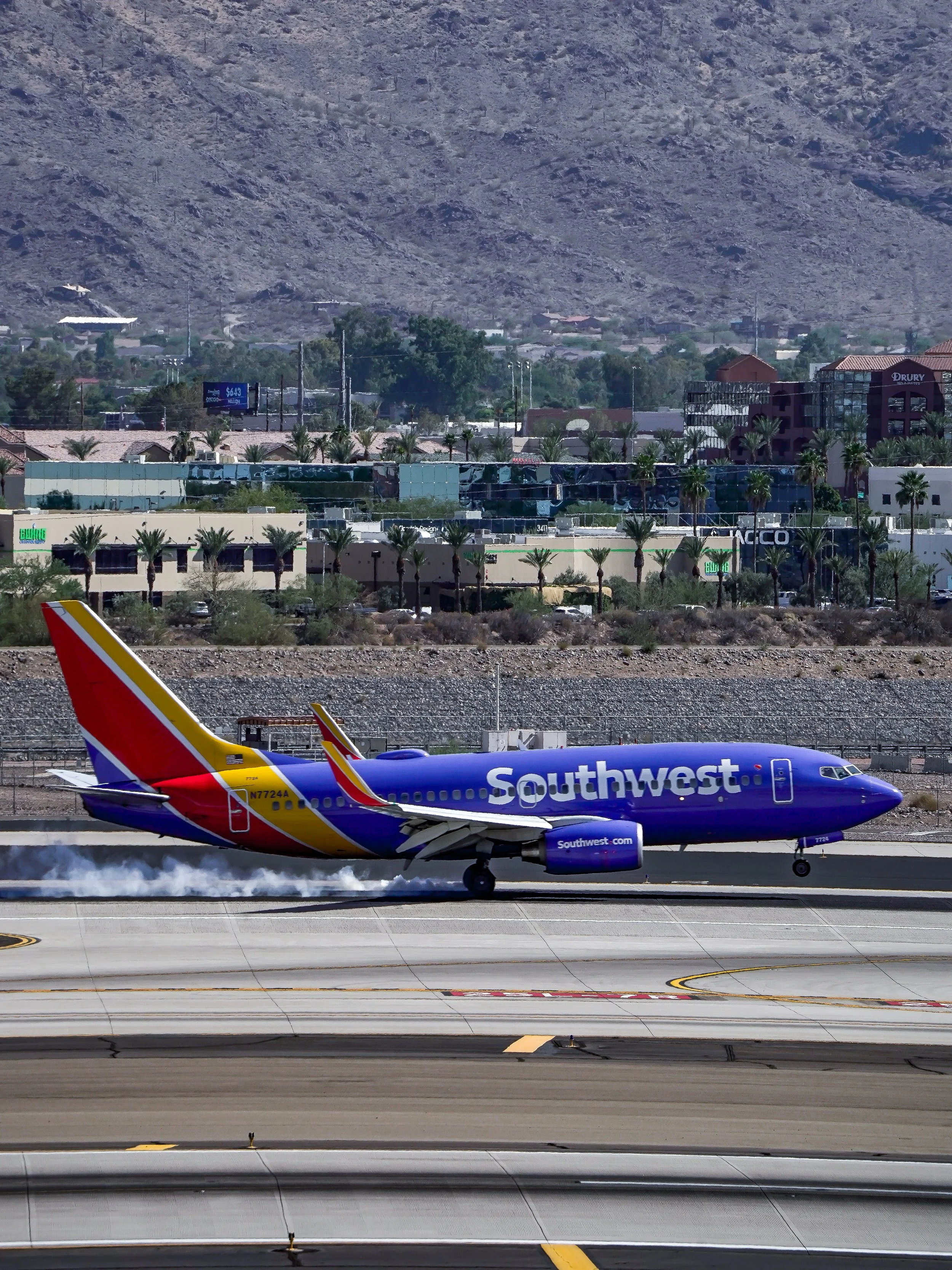 A Southwest Airlines airplane landing on a runway, creating smoke from the tires, with a desert landscape and a cityscape with palm trees and buildings in the background.