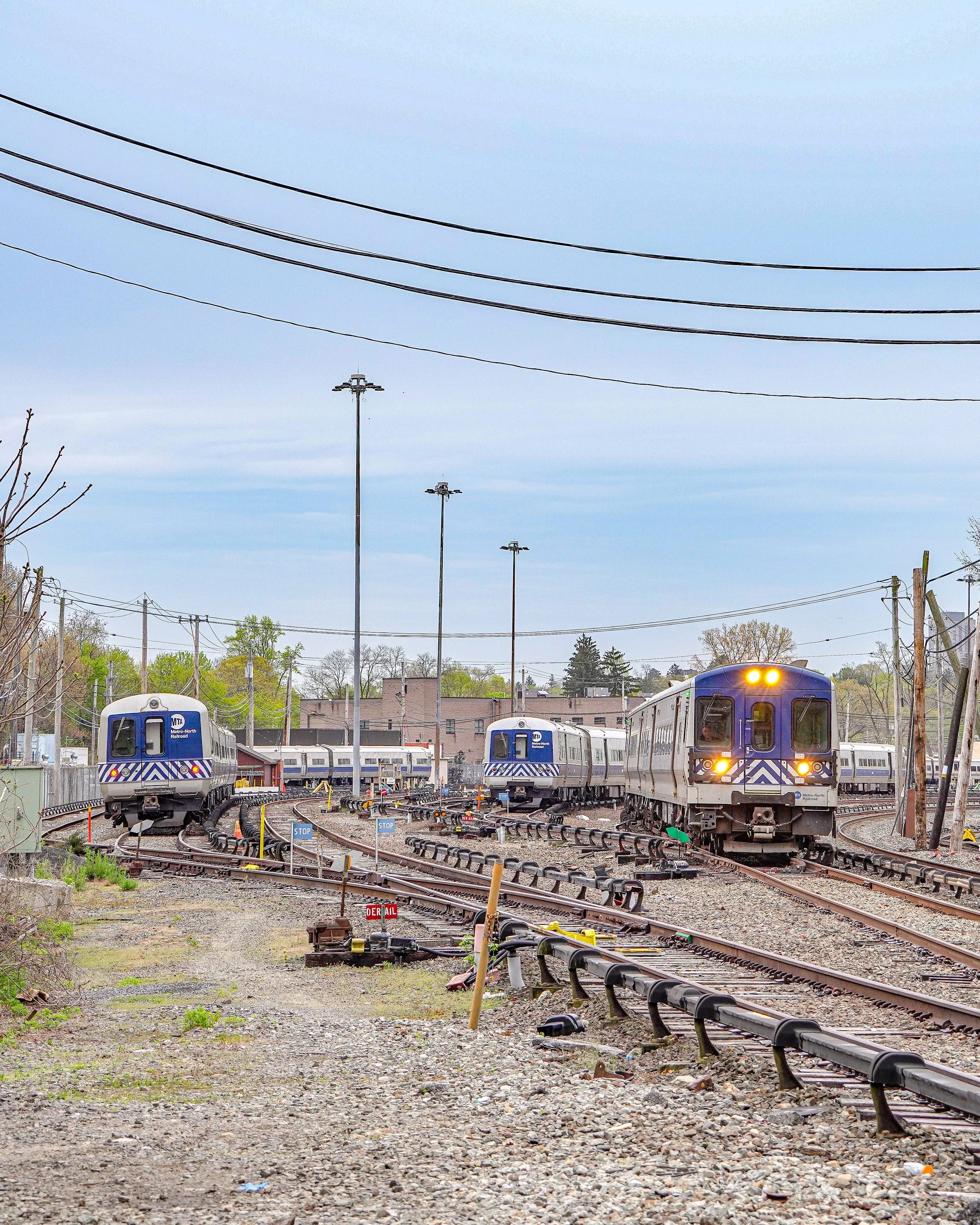 Multiple subway trains on tracks in a train yard with utility poles and wires, some buildings in the background, and a mostly cloudy sky.
