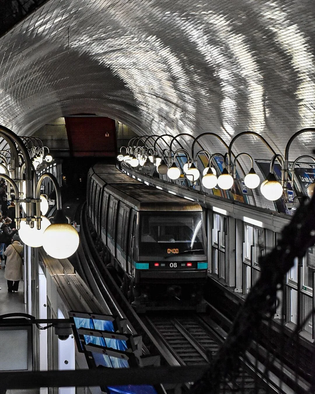 A subway train arriving at an underground station with a curved, tiled ceiling reflecting the light. The platform is lit by vintage-style globe lamps, and passengers are waiting.