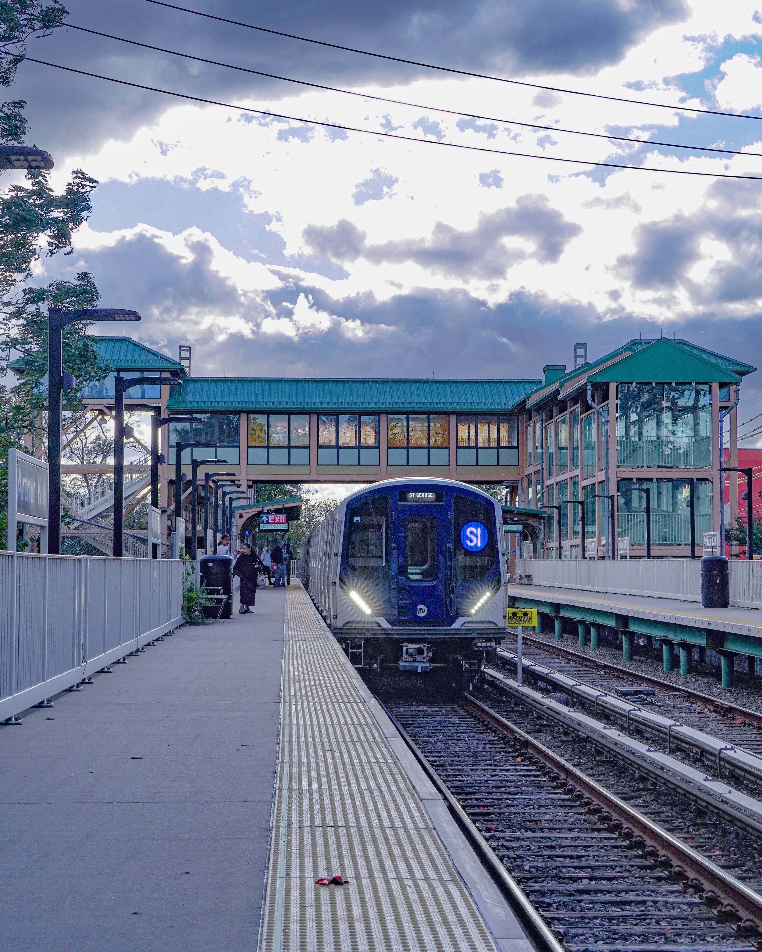 A light rail train approaching a station platform with pedestrians waiting, a blue ADA sign, and a sky with clouds.