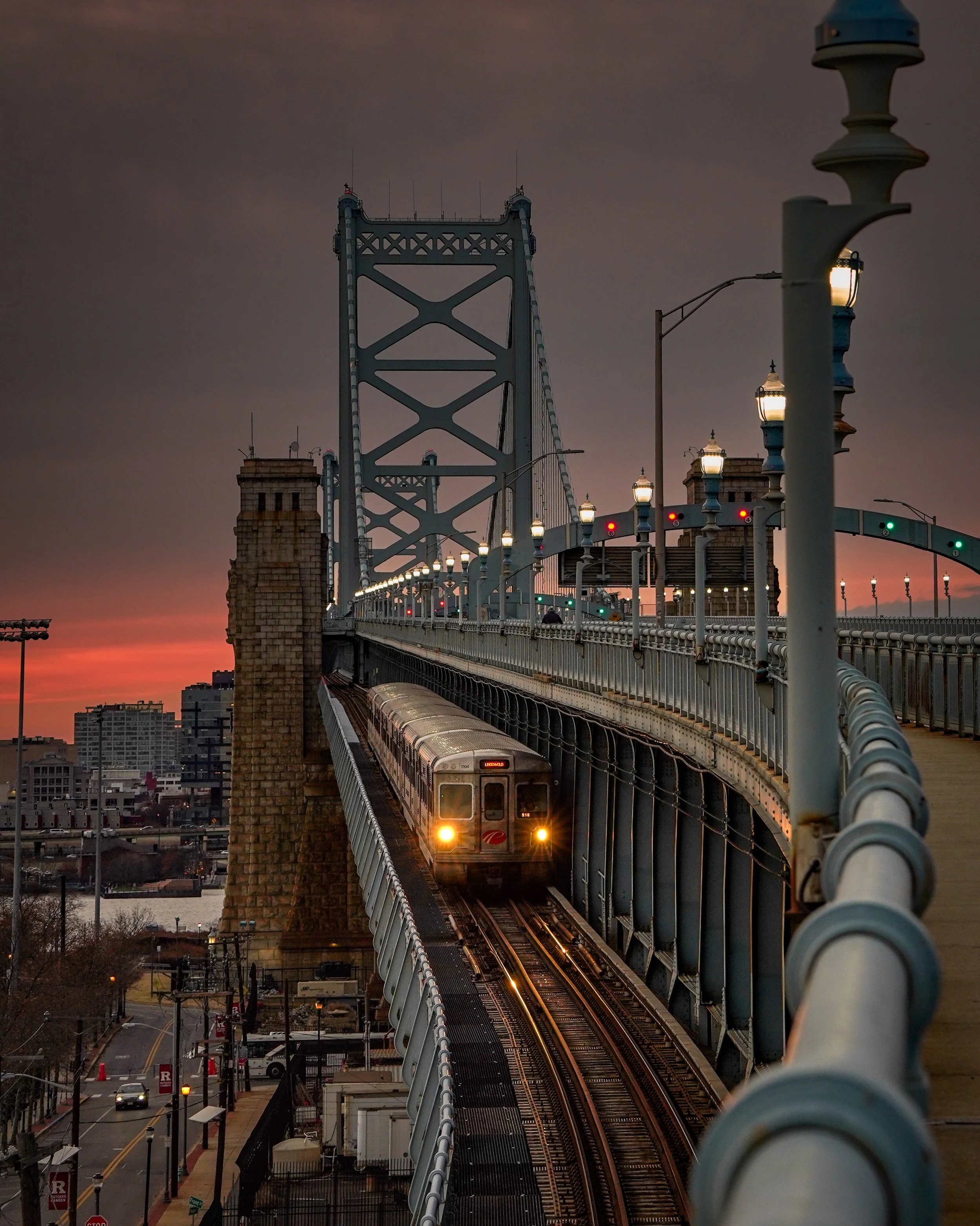 A PATCO train on the Benjamin Franklin Bridge during sunset in Philadelphia, with city buildings in the background.