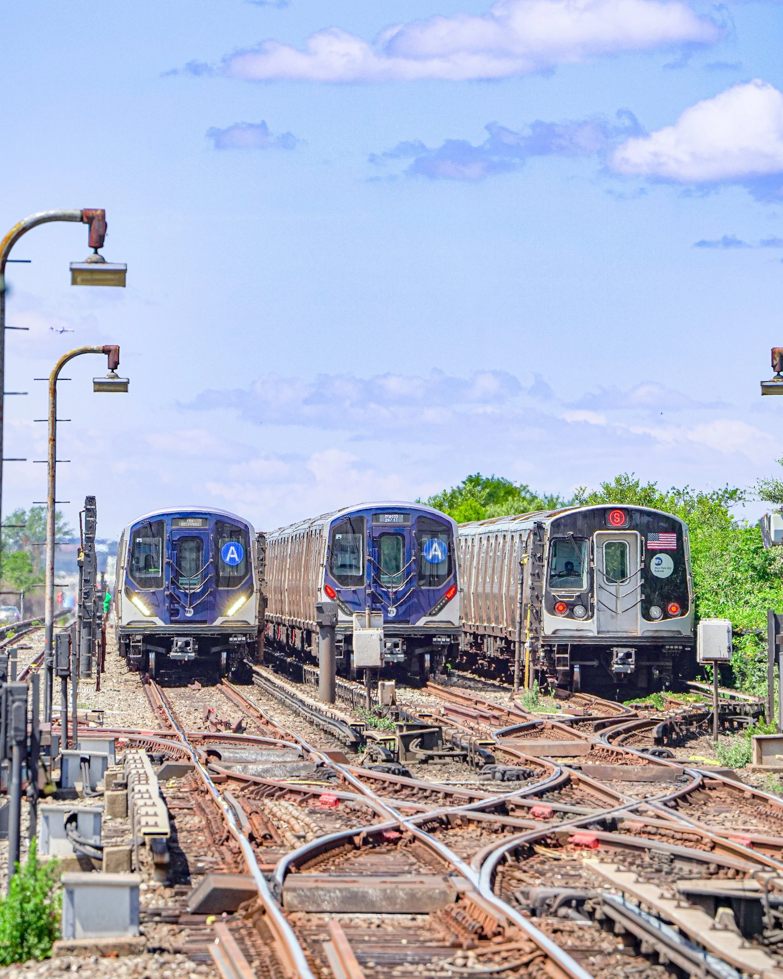Three metro trains on parallel tracks with a background of trees and a sky with clouds.