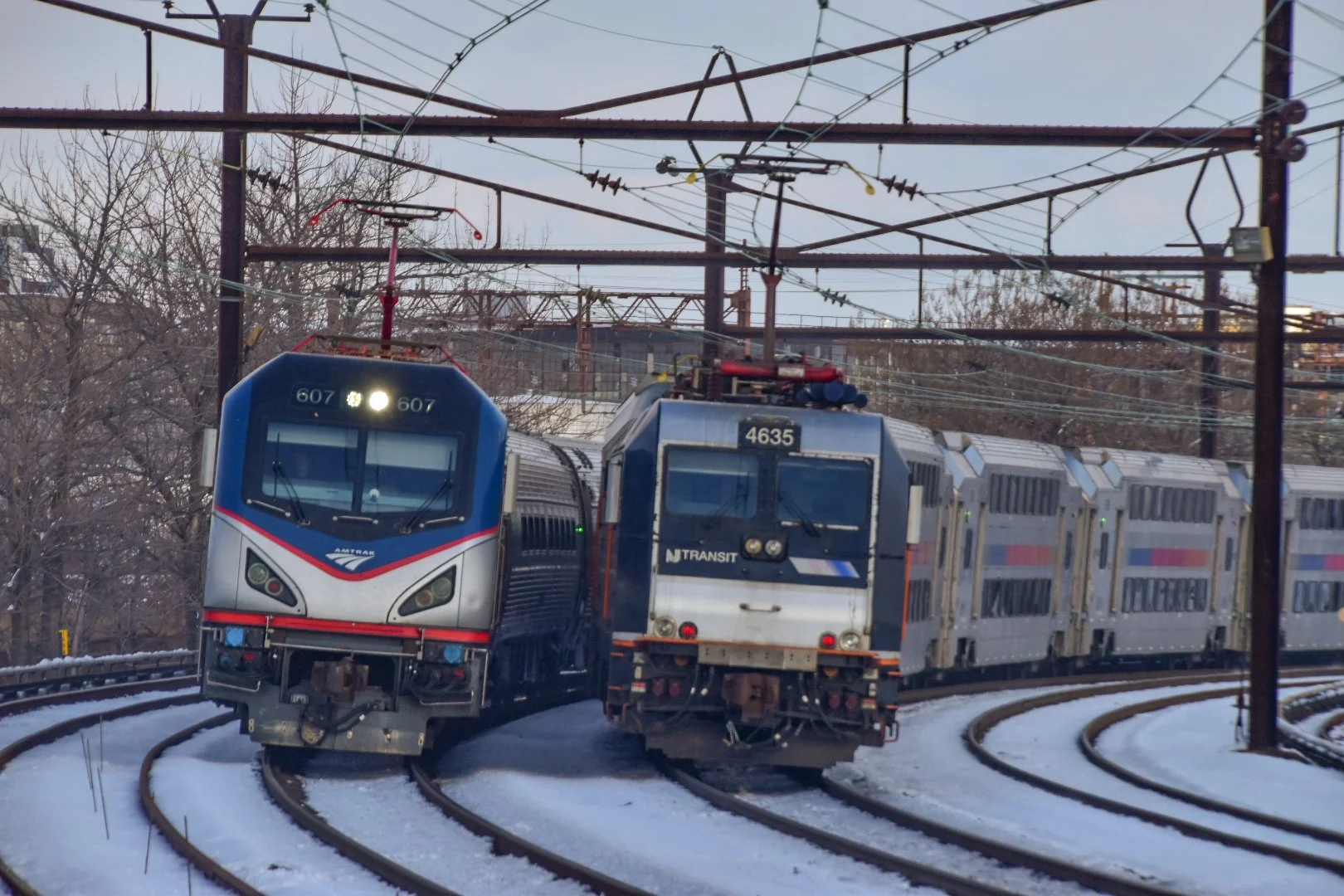 A New Jersey Transit train diverging from another train on snowy tracks, with trees and utility lines overhead.