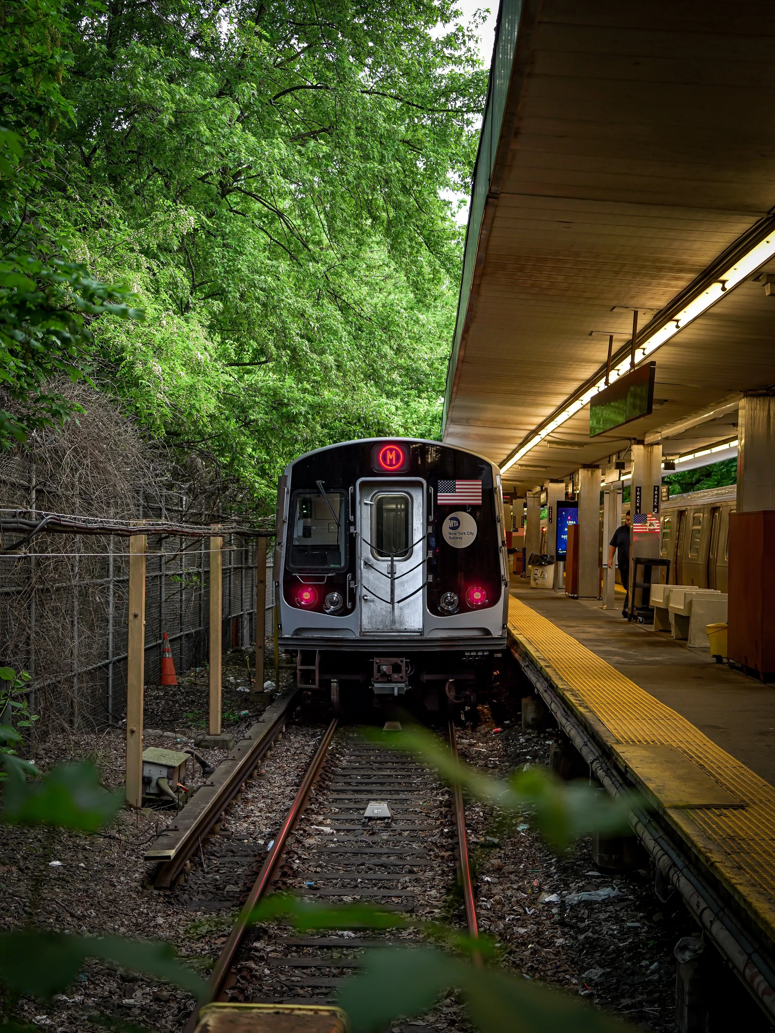 Subway train at a station platform with a green leafy background and a partially visible ceiling, with platform lighting and a person walking in the background.